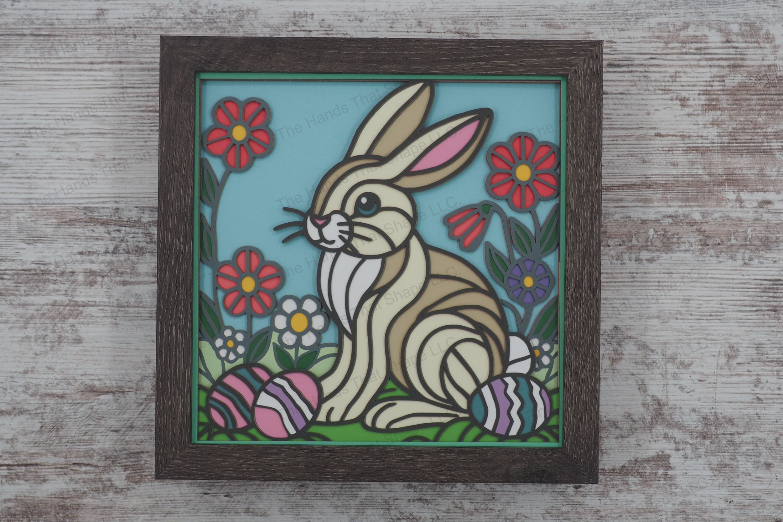 A charming framed layered paper shadowbox featuring a bunny sitting in a grassy meadow surrounded by colorful Easter eggs and blooming spring flowers.