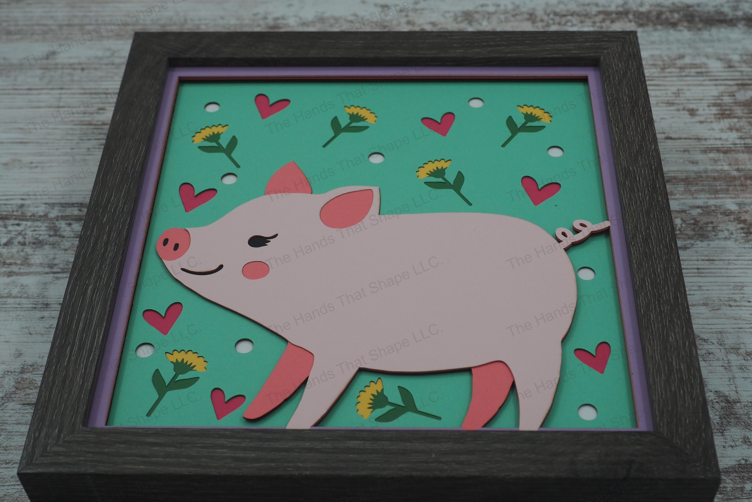 This layered paper shadowbox features a folksy piglet, surrounded by charming flowers, dots, and hearts, perfect for adding whimsical charm to any room.