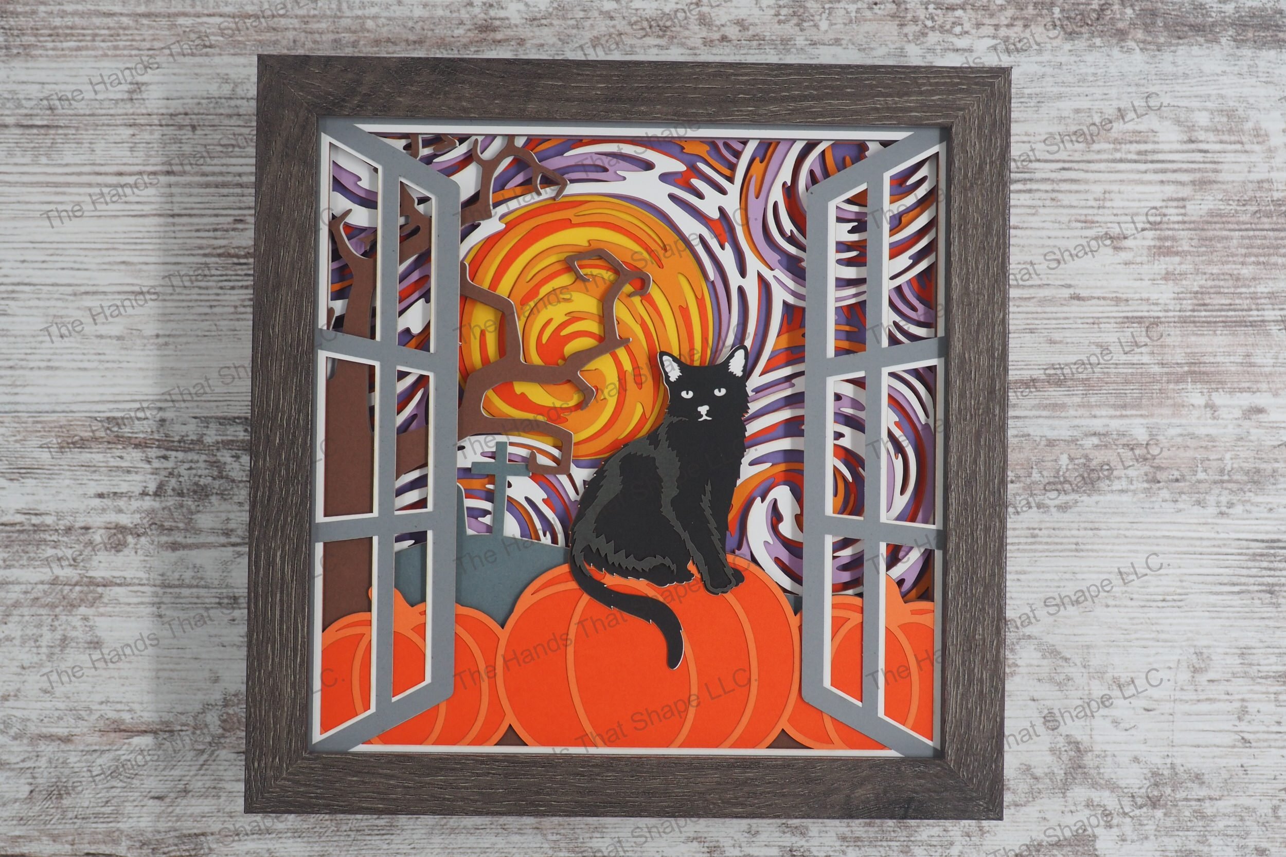 A layered paper shadowbox depicting a Halloween scene through a window with a black cat perched on a pumpkin under a swirling, colorful sky on a full moon night