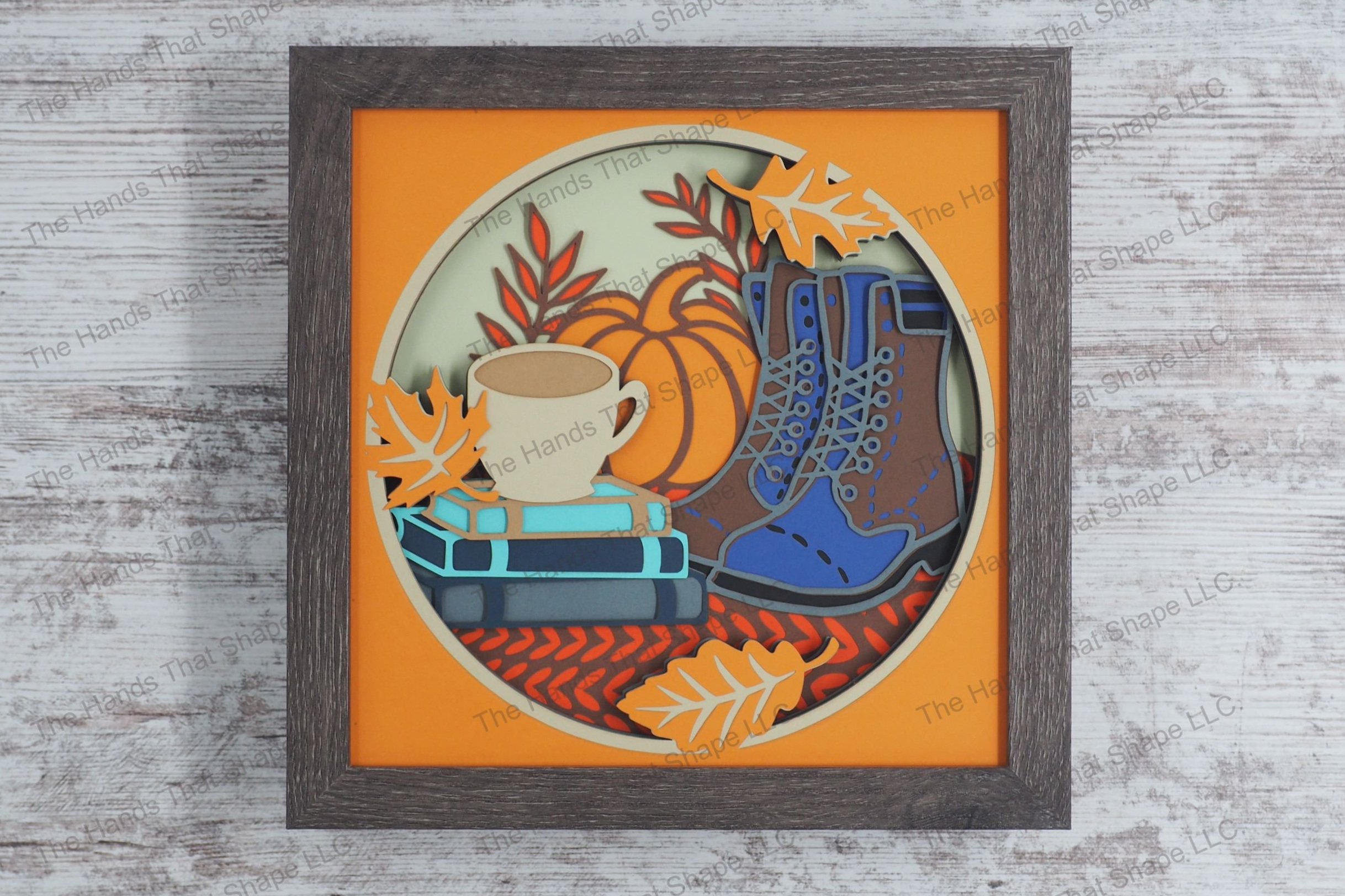 A cozy autumn-themed layered paper shadowbox depicting a stack of books with a coffee cup on top, a pair of boots and a large pumpkin, and fall leaves.