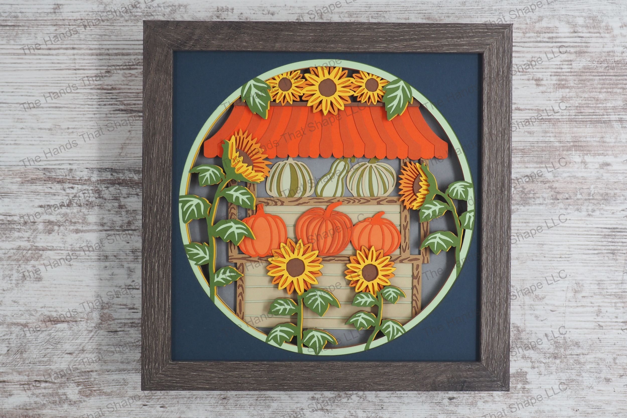 A vibrant framed layered paper shadowbox depicting a market stand filled with pumpkins and surrounded by sunflowers, crafted from 15 layers of cardstock.