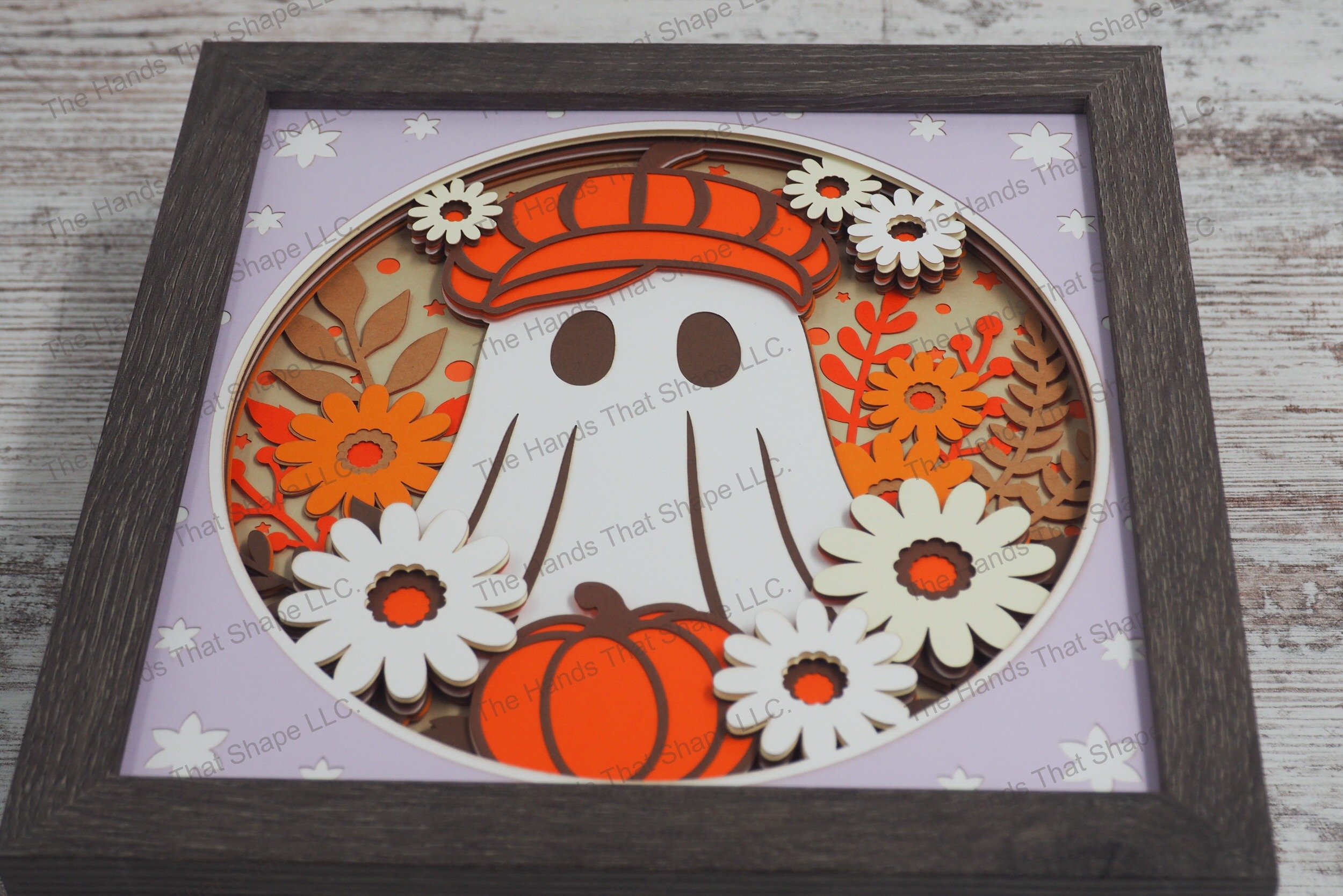 A framed playful layered paper shadowbox depicting a friendly ghost adorned with a hat, surrounded by vibrant fall flowers and a pumpkin.