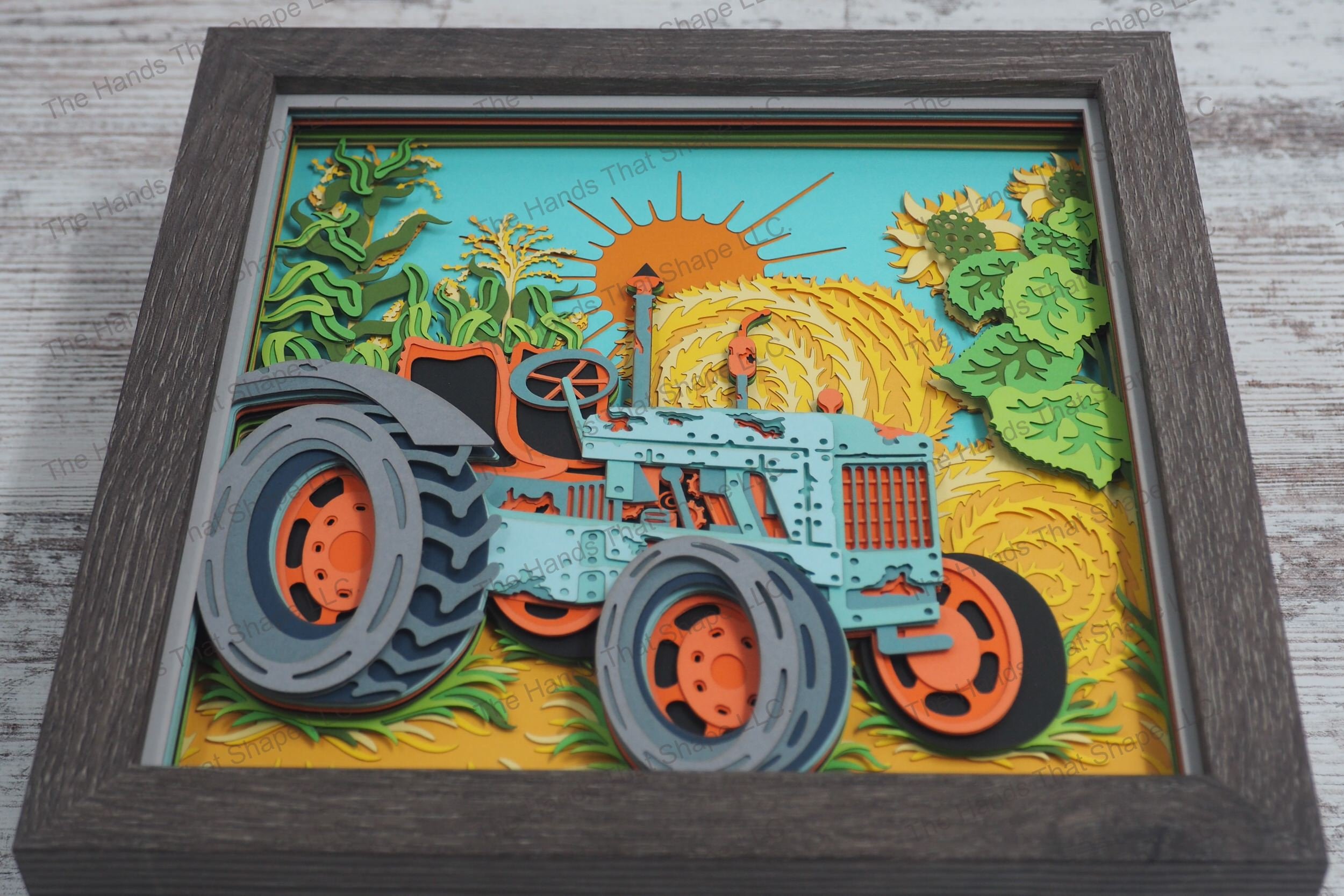 A detailed layered paper framed shadowbox depicting an old tractor surrounded by corn stalks, sunflowers, and hay bales under a setting sun.