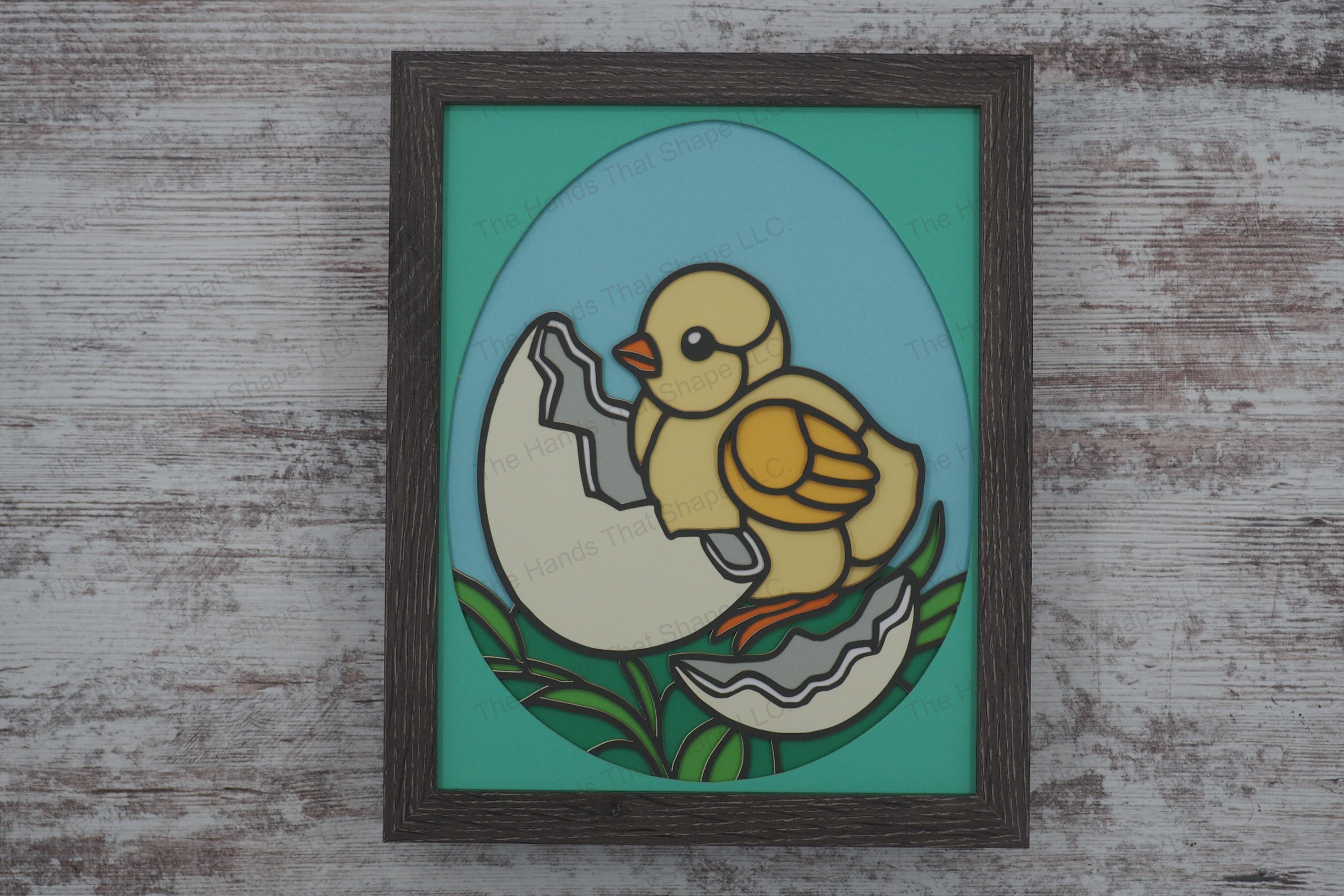 This charming layered paper shadowbox features a freshly hatched chick beside its eggshell, set against a grassy backdrop, all encased in an egg-shaped border.