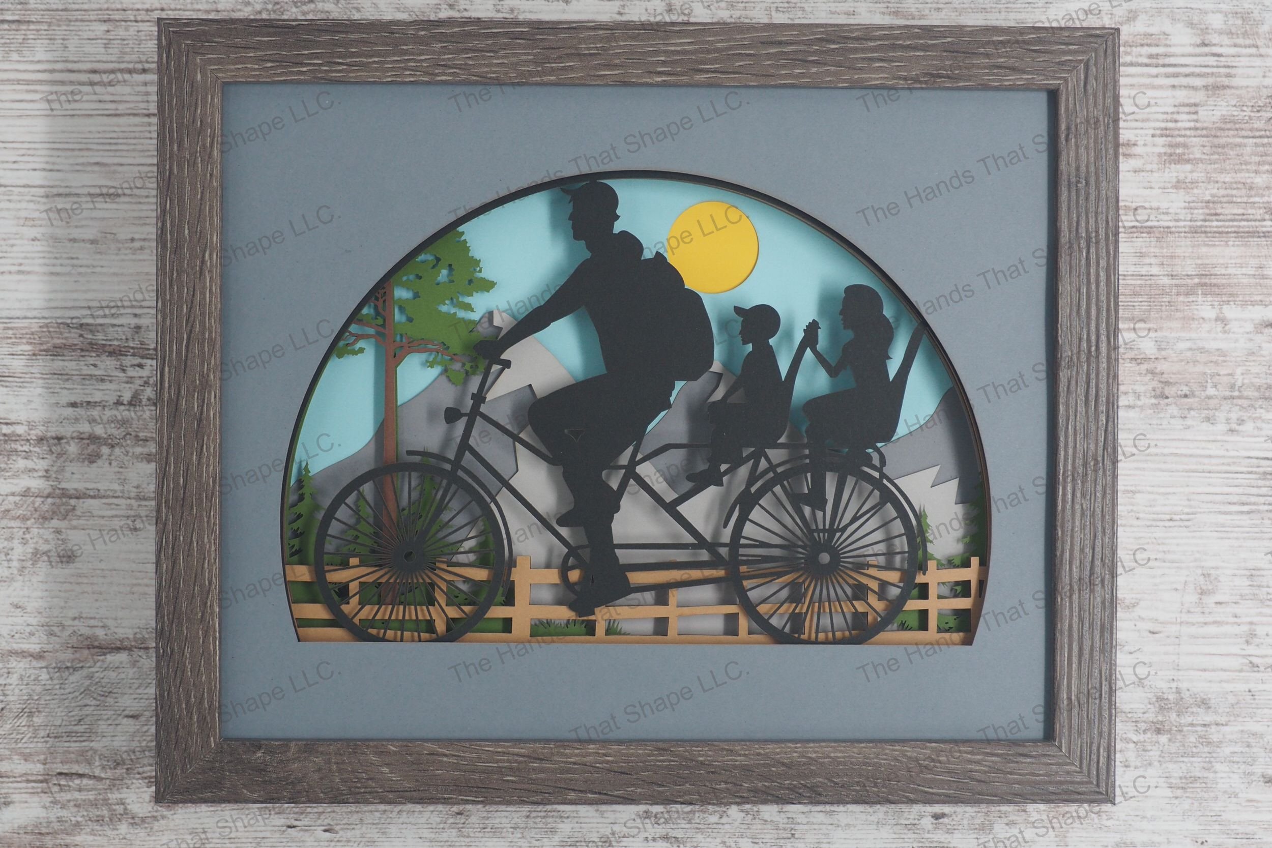A layered paper shadowbox depicting a family bike ride featuring a father and two children on a bicycle against a backdrop of a mountain range.