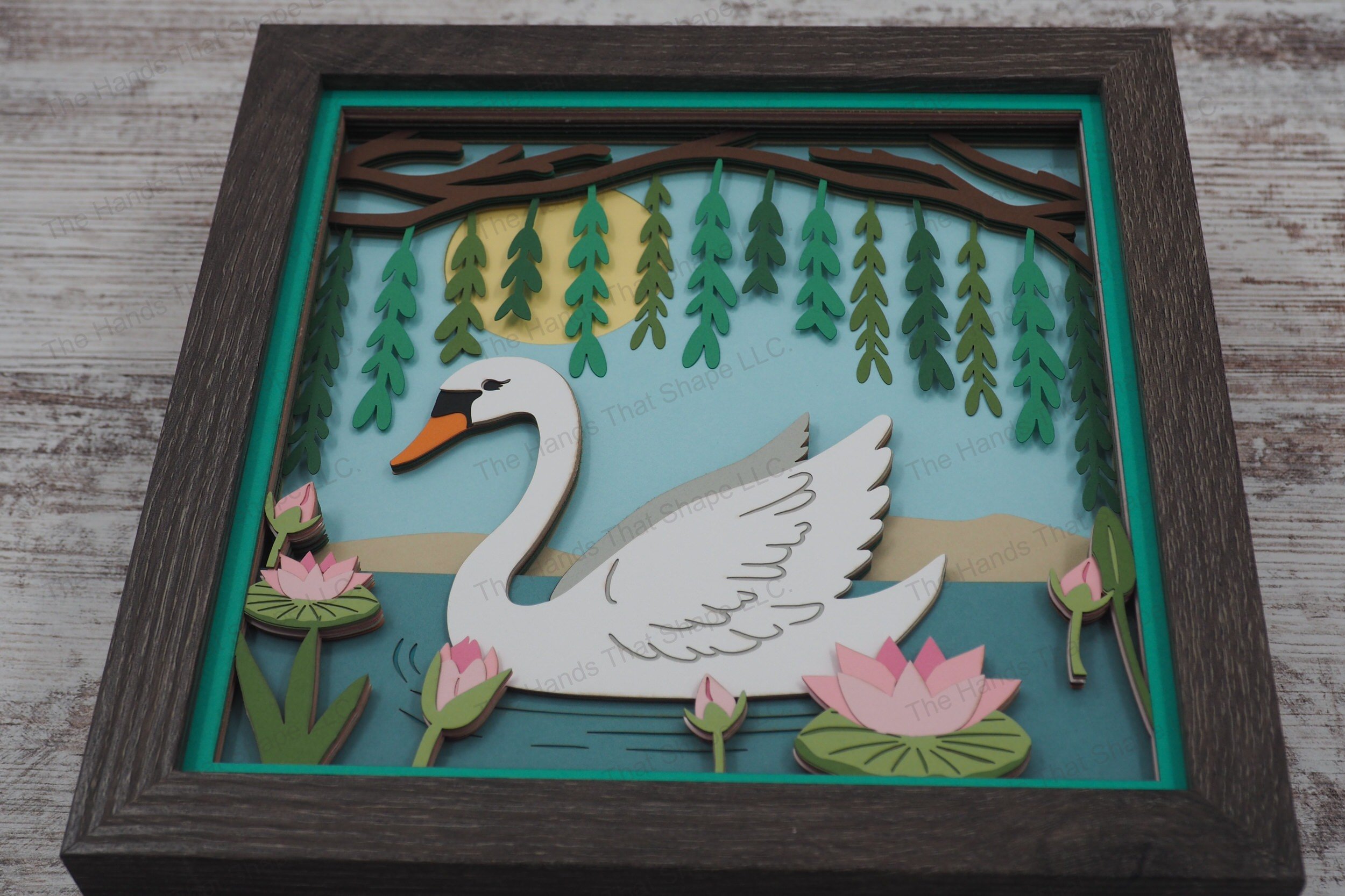 A layered paper shadowbox featuring a swan swimming beneath a willow tree, surrounded by lily pads and flowers, creating a serene and captivating 3D scene.
