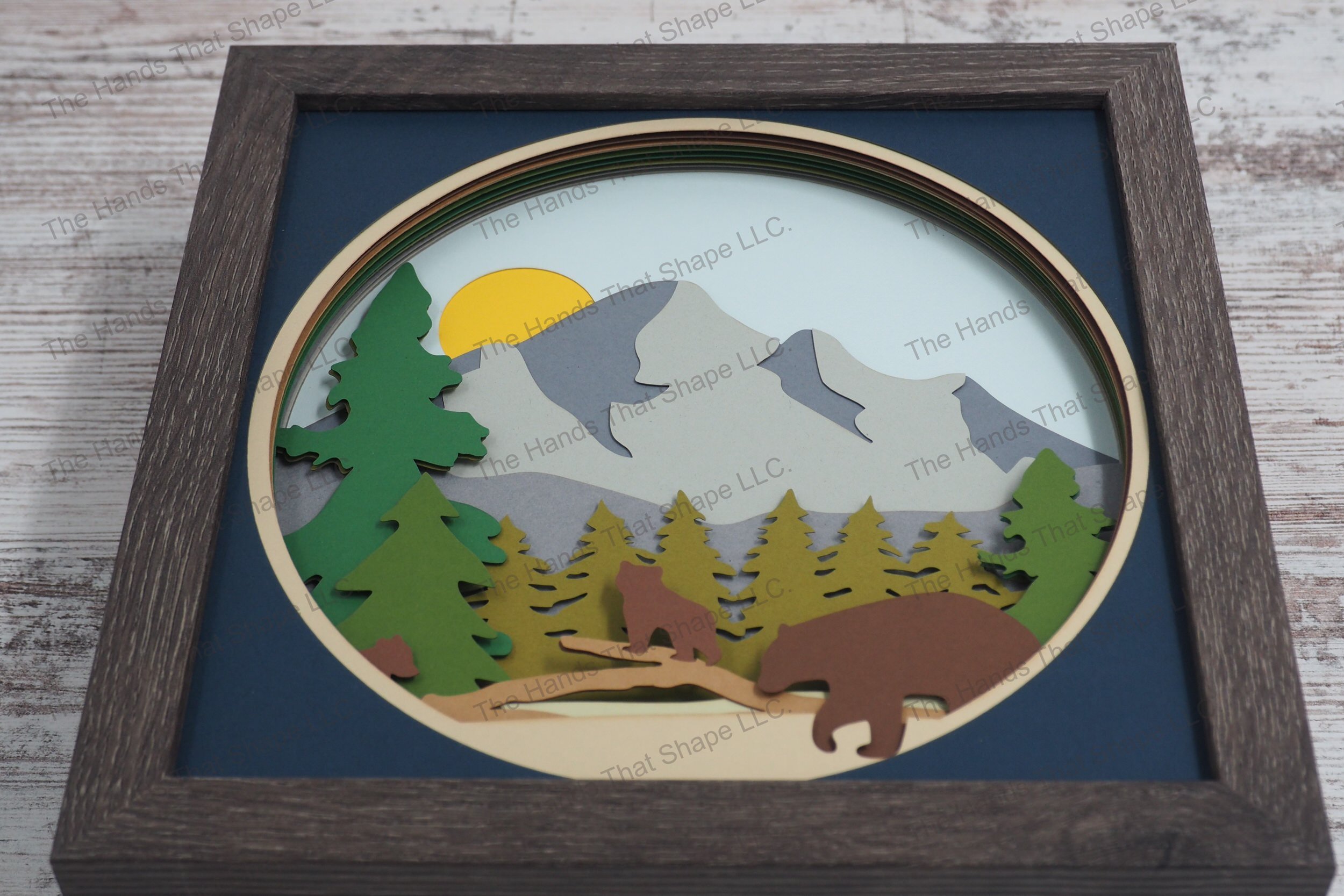 A charming depiction of a bear family interacting in a mountain forest, this layered paper shadowbox captures mama bear and her cubs among pine trees.