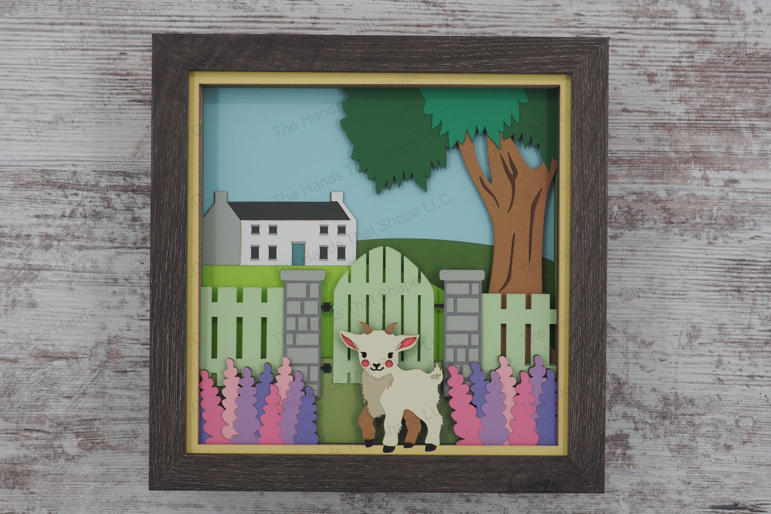 This layered paper art shadowbox features a goat standing in a flower garden, beside a rustic fence framed by blooming hyacinths and a countryside house.