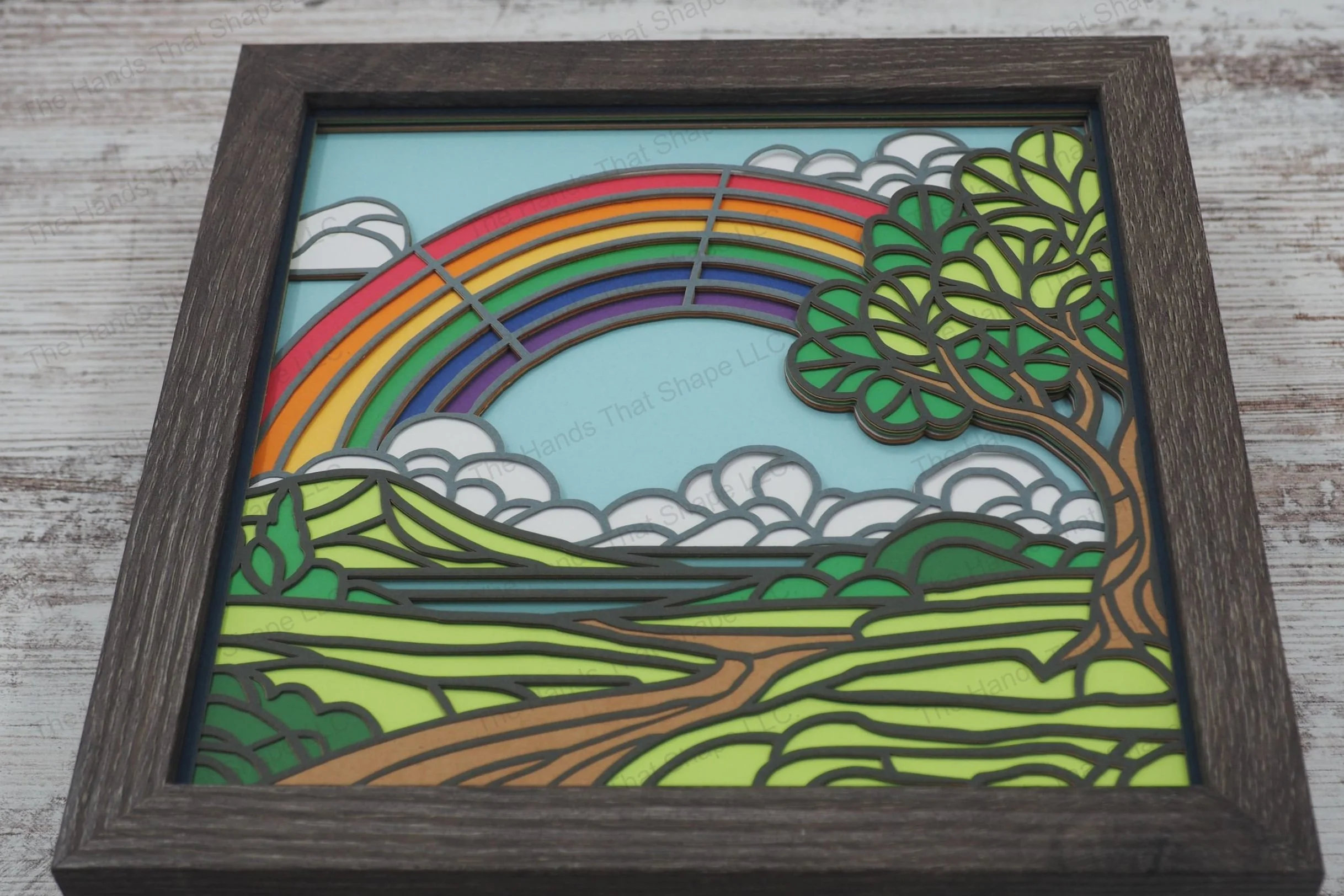This layered paper shadowbox features a vibrant rainbow arching over a picturesque countryside with rolling hills, and a winding path to a distant lake.