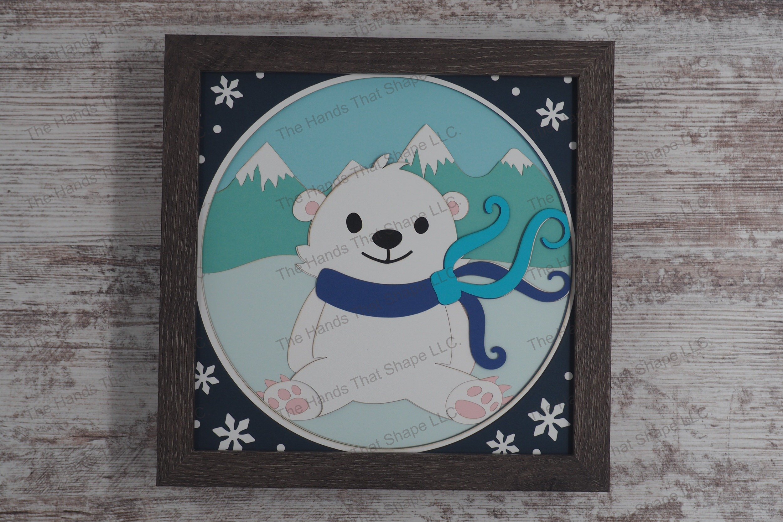 A charming layered paper shadowbox depicting a polar bear cub wearing a colorful scarf, set against a backdrop of snowy mountains and bordered with snowflakes.