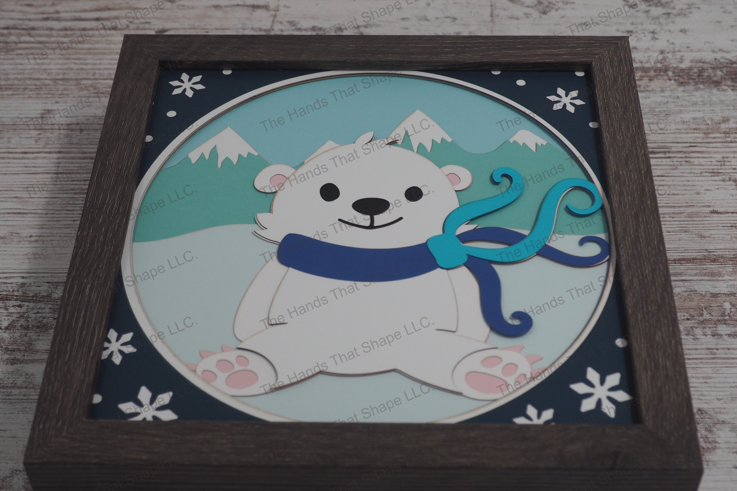 A charming layered paper shadowbox depicting a polar bear cub wearing a colorful scarf, set against a backdrop of snowy mountains and bordered with snowflakes.
