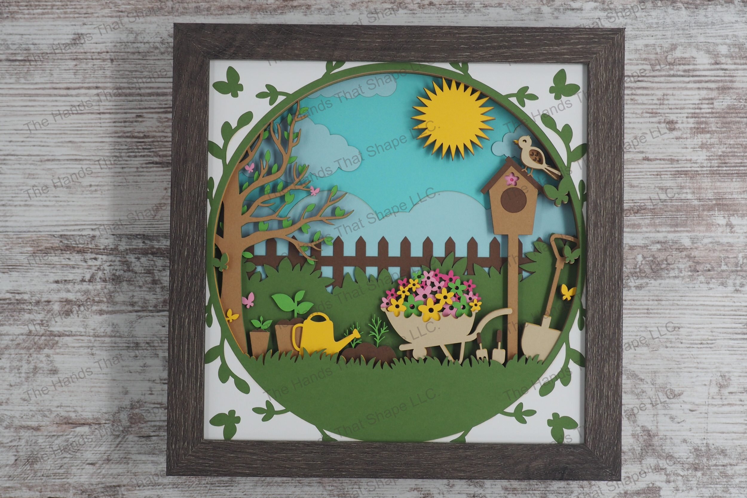 A detailed layered paper shadowbox depicting a lush backyard garden scene with a wheelbarrow full of flowers, gardening tools, butterflies, and a sunlit sky.