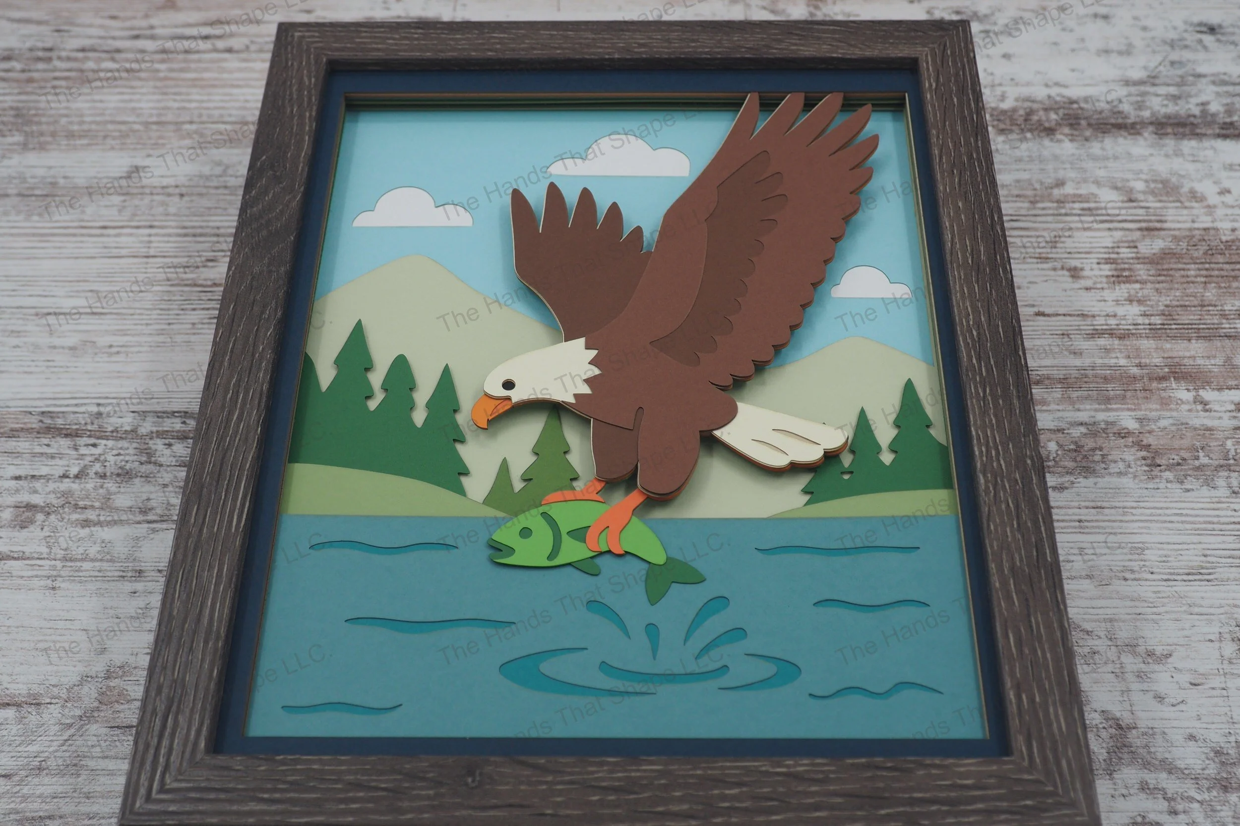 A handcrafted 3D layered paper shadowbox featuring a bald eagle swooping over a lake to catch a fish, set against layered hills, pine trees, and mountains.