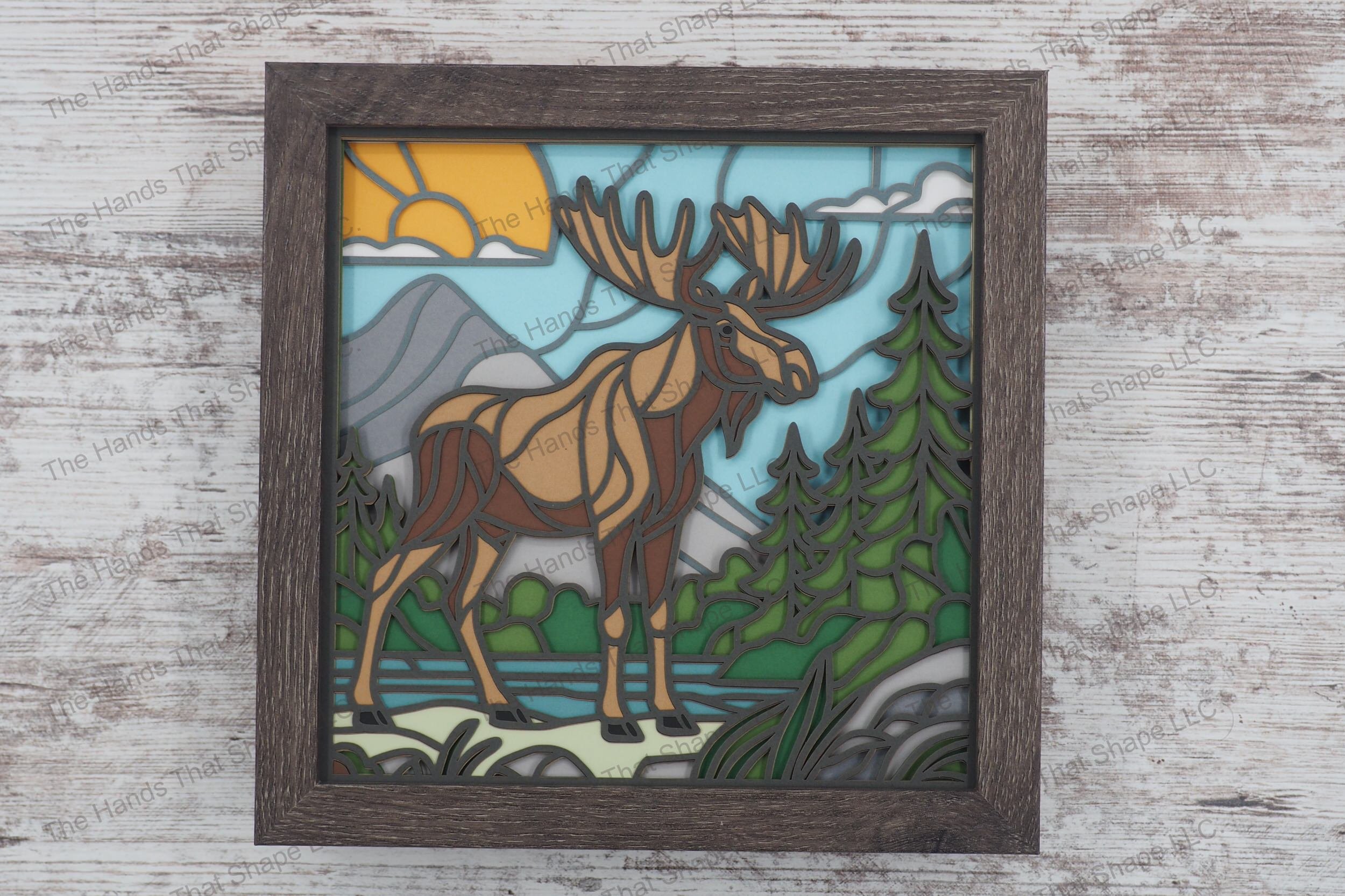 Explore the beauty of the wilderness with this layered paper shadowbox, featuring a moose at a water's edge, set against a backdrop of pine trees and mountains.