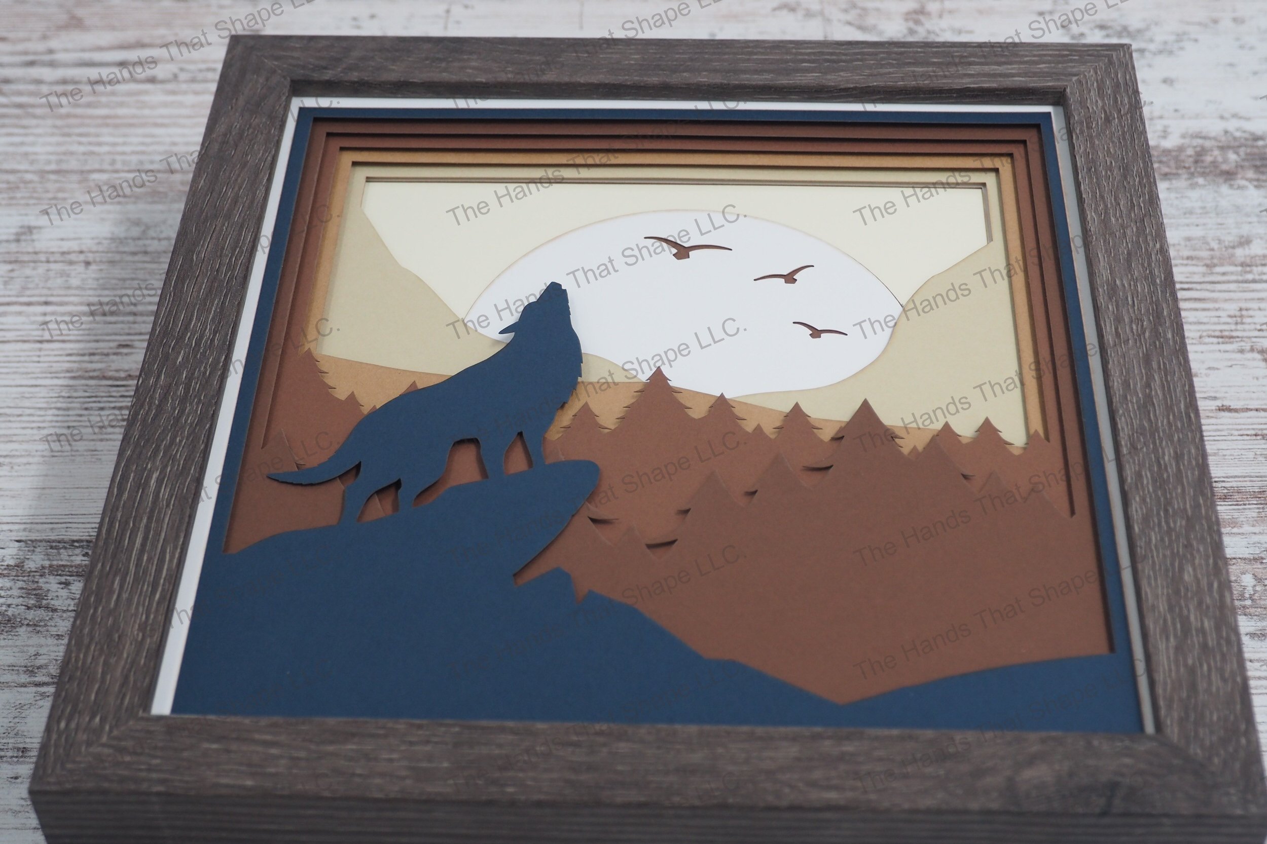 A framed layered paper shadowbox depicting a lone wolf howling at a full moon from a rocky ledge, with a backdrop of pine trees, rolling hills, and mountains.