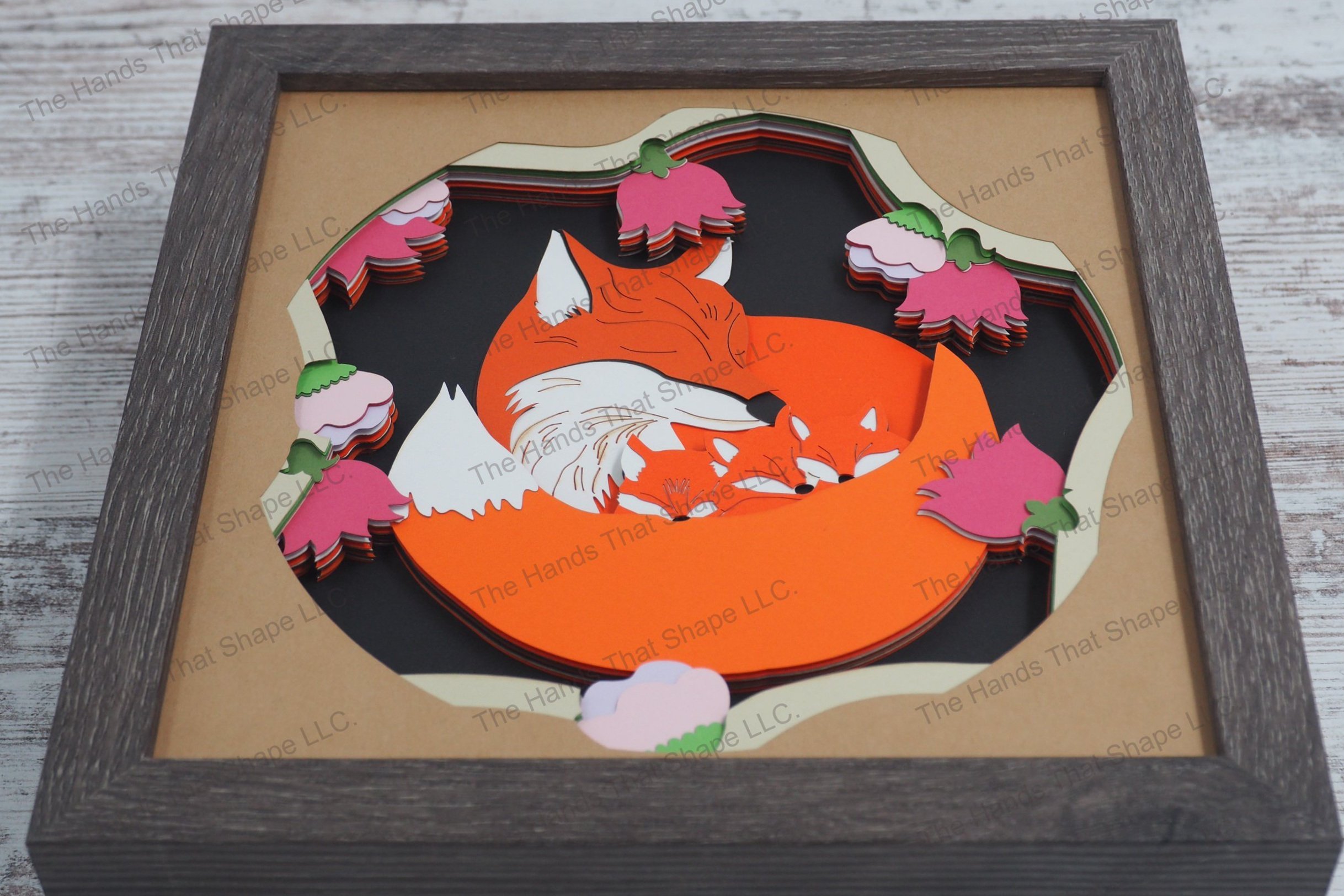 A captivating layered paper shadowbox depicting a mama fox tenderly curled around her three kits among vibrant flower blossoms in a gray MDF frame.