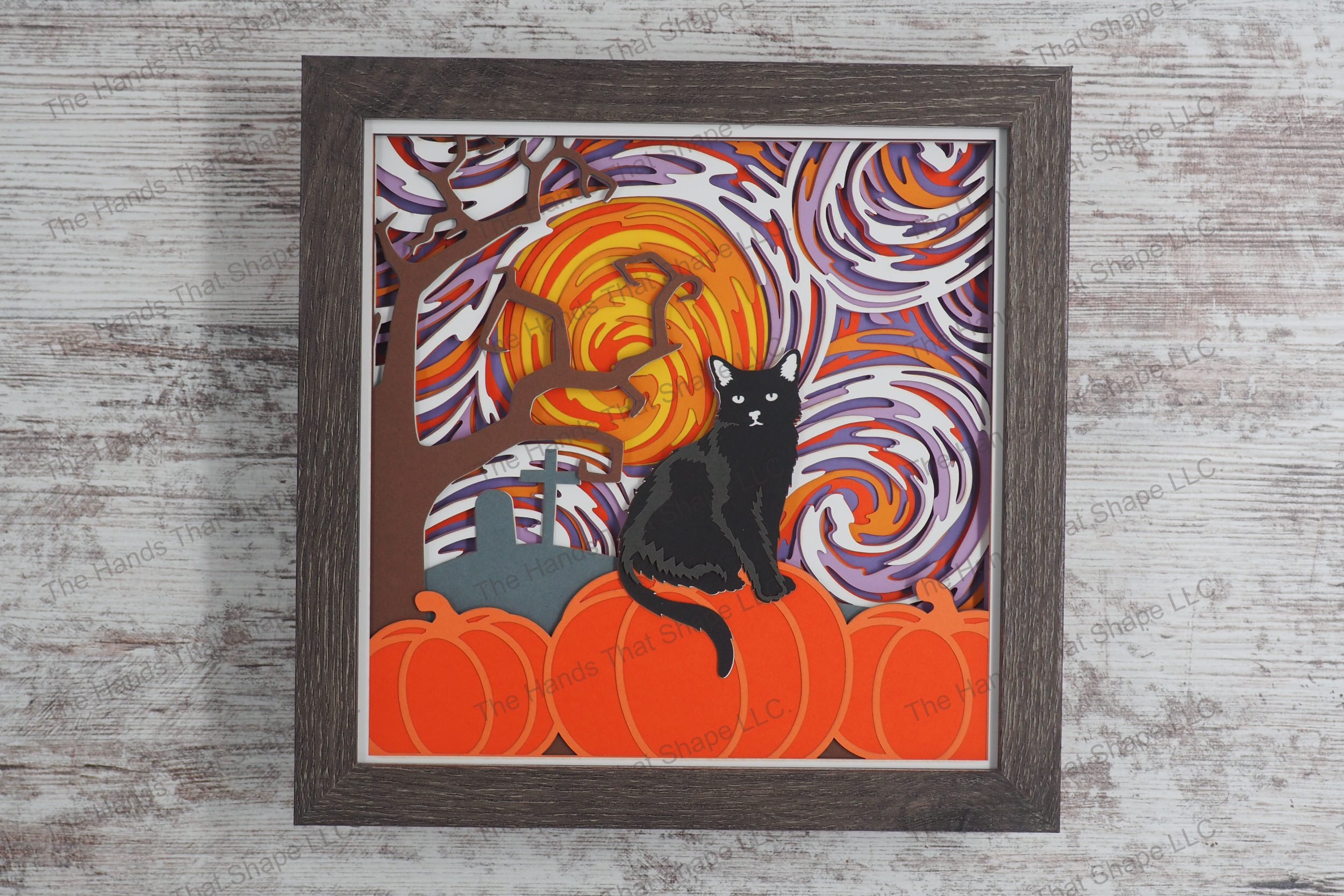 A framed layered paper shadowbox depicting a Halloween scene with a black cat perched on a pumpkin under a swirling, colorful sky on a full moon night.