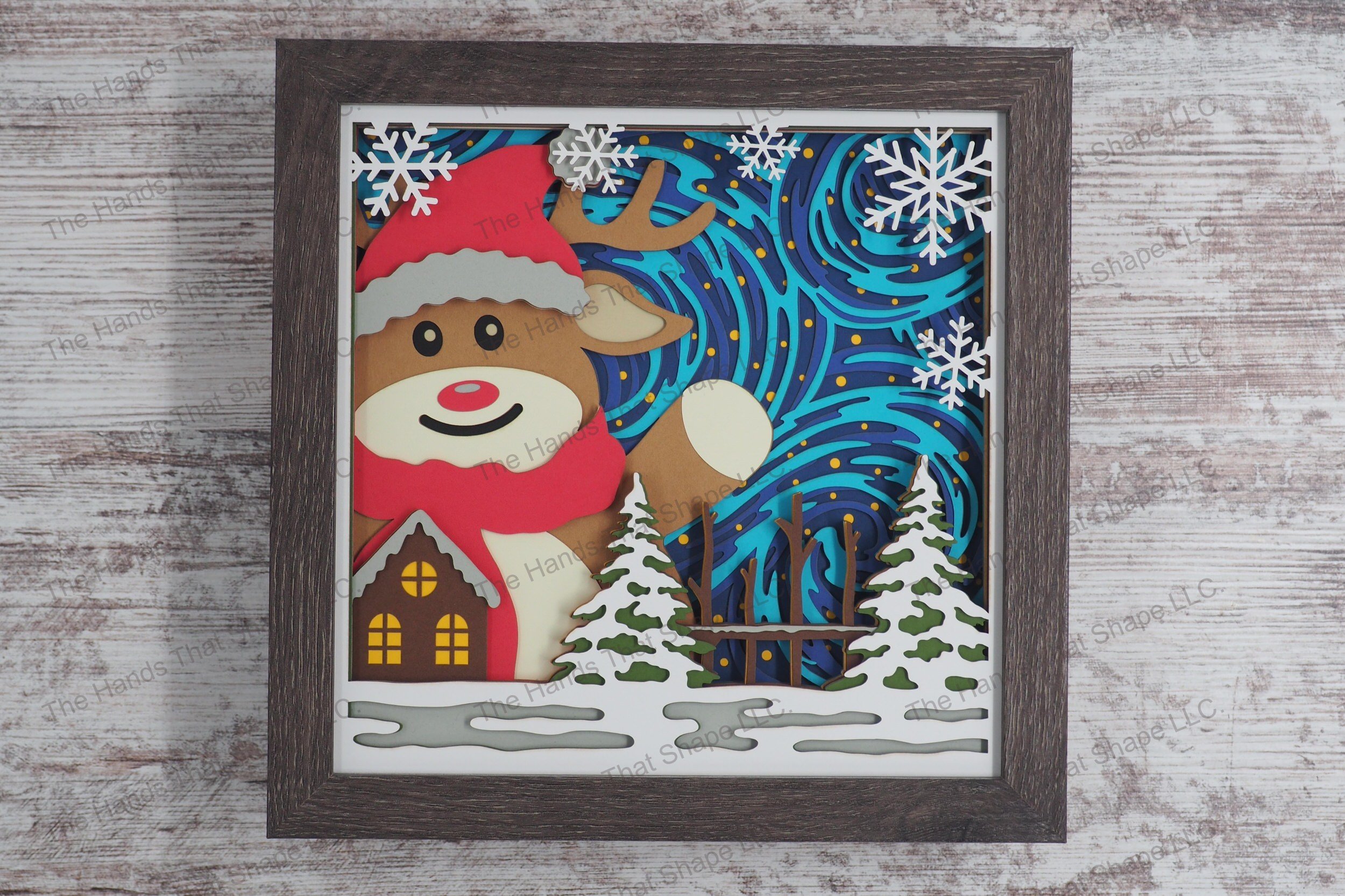 A layered paper shadowbox depicting an oversized festive reindeer waving joyfully beside a snow-covered cabin, with wintery swirls and stars as a backdrop.