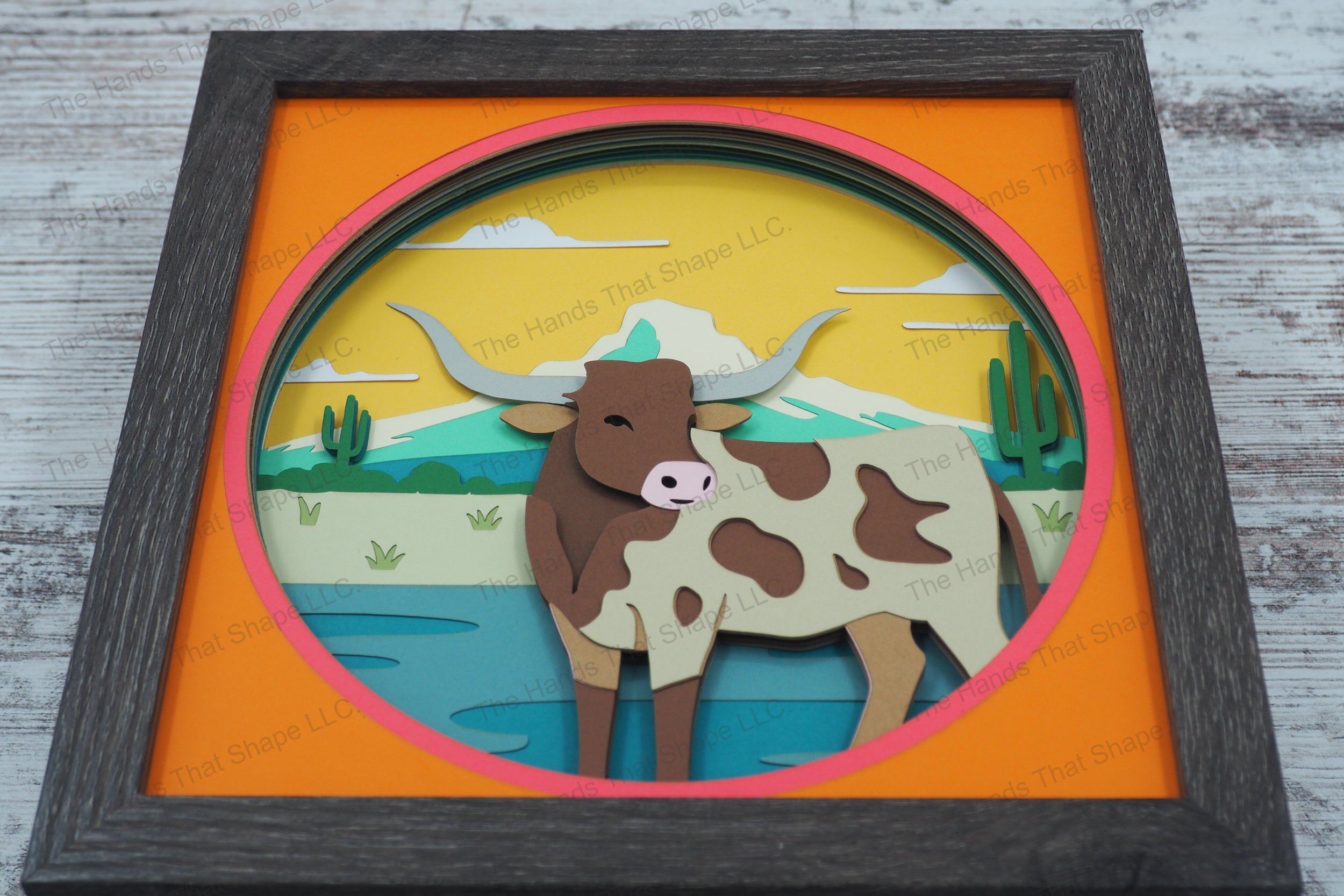 A detailed 3D layered paper shadowbox showcasing a Texas Longhorn standing in water with a Southwestern backdrop of cactuses, hills, and mountains.