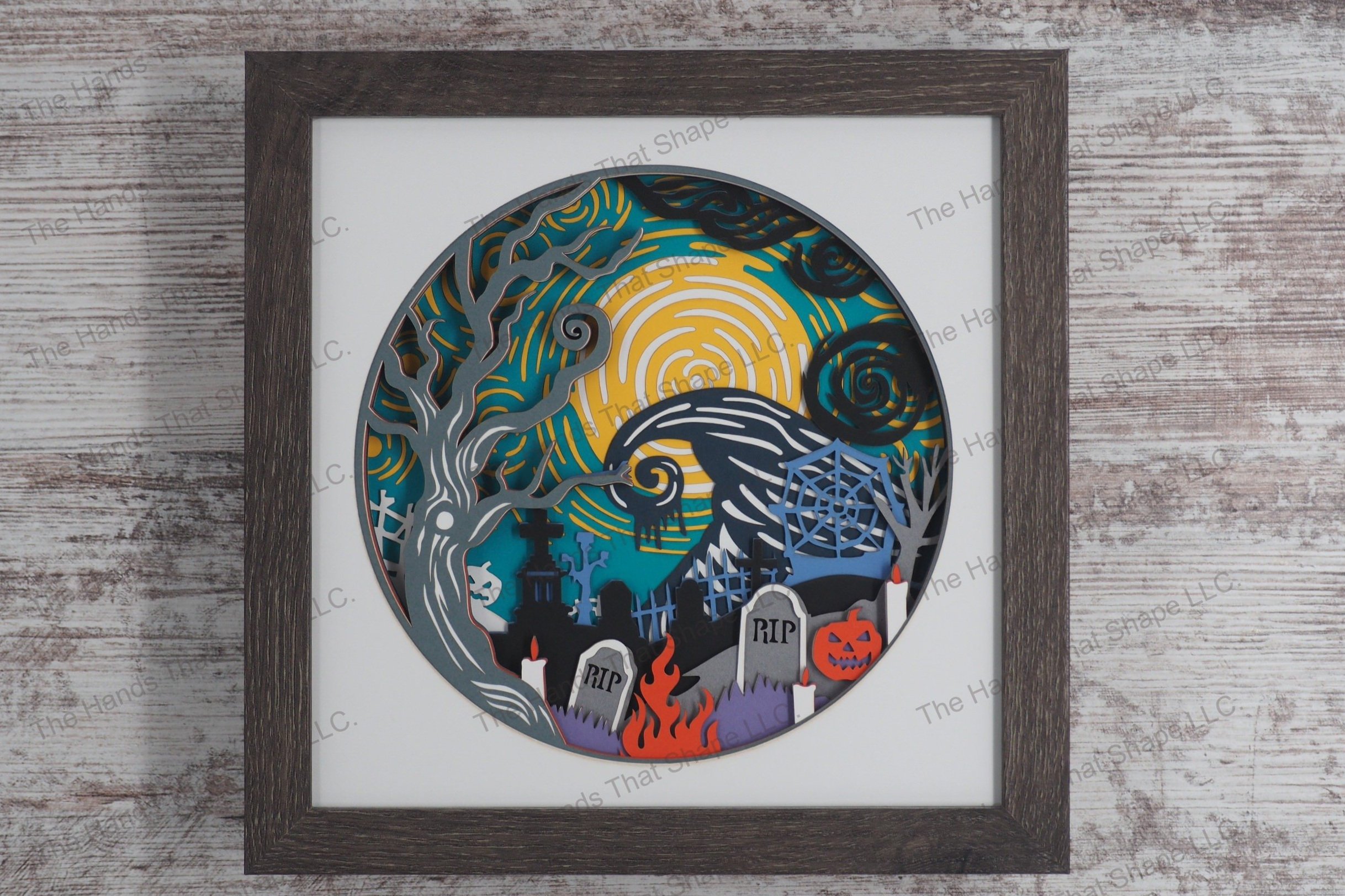 A captivating layered paper framed shadowbox depicting a spooky Halloween cemetery scene with aged tombstones, an ancient tree and a witch's hat-shaped hill.