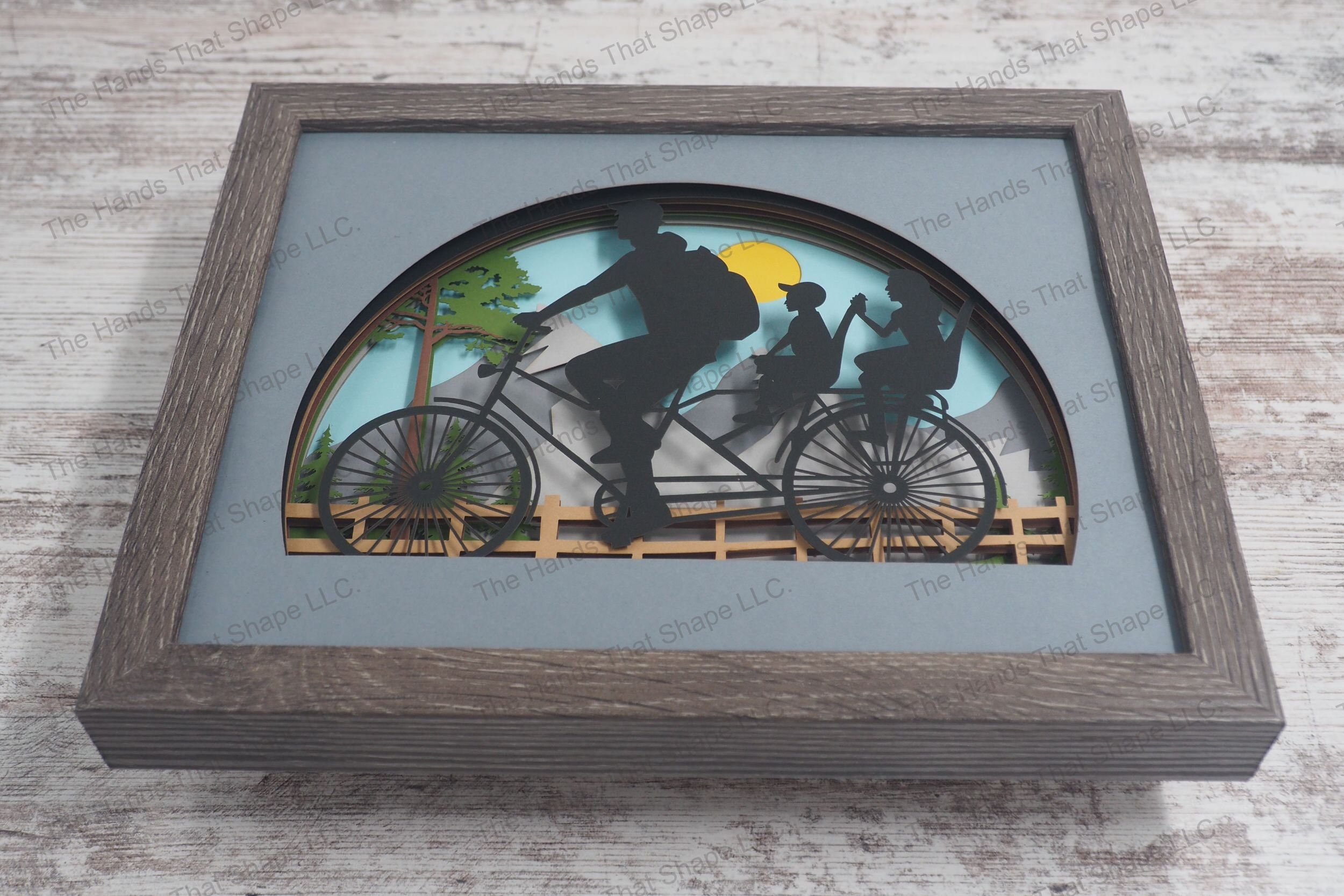 A layered paper shadowbox depicting a family bike ride featuring a father and two children on a bicycle against a backdrop of a mountain range.