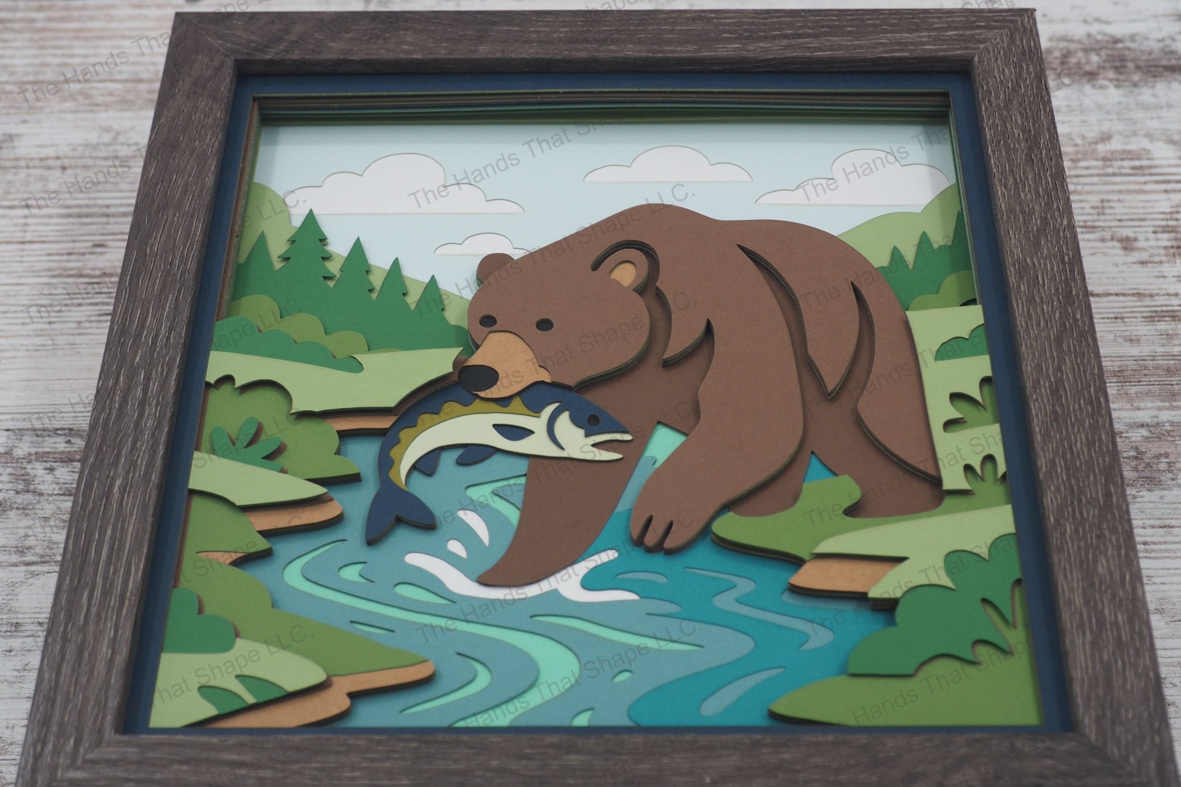 A dramatic 3D layered paper shadowbox featuring a grizzly bear standing in a stream with a fish in its mouth, framed by lush greenery, trees, and a cloudy sky.