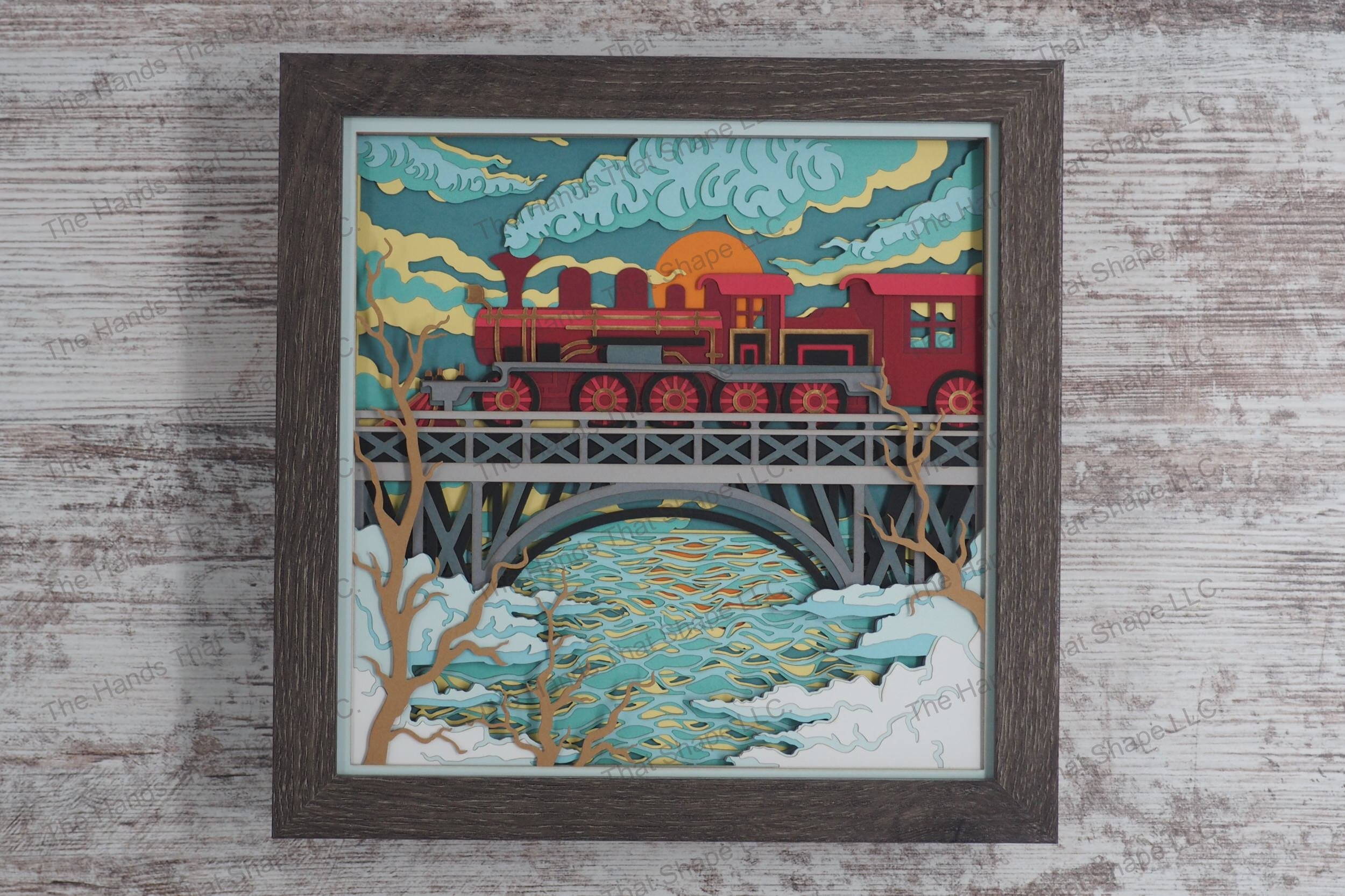 A detailed layered paper framed shadowbox depicting a classic steam train billowing smoke as it crosses a high bridge over a river, with a setting sun.