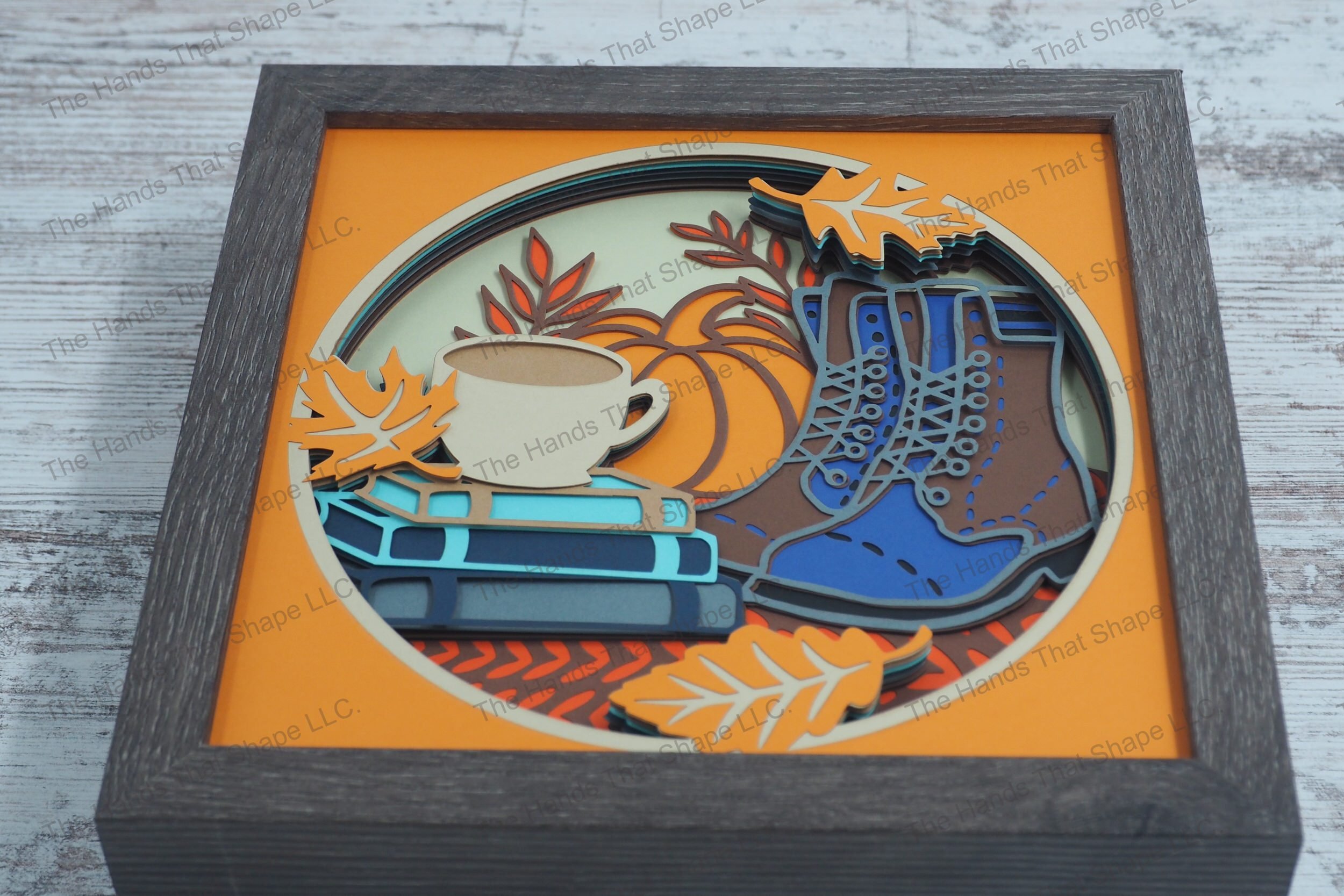 A cozy autumn-themed layered paper shadowbox depicting a stack of books with a coffee cup on top, a pair of boots and a large pumpkin, and fall leaves.