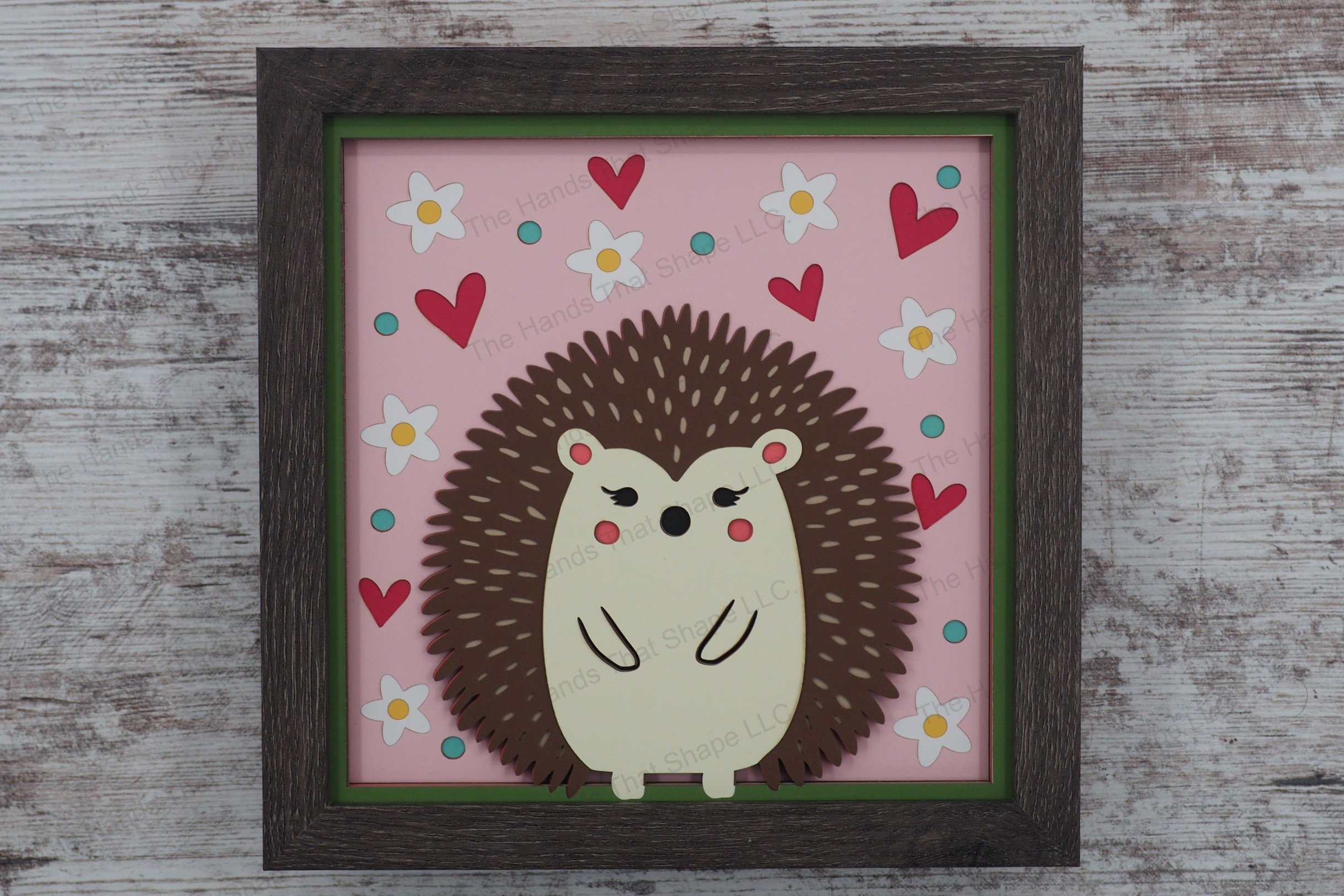 This layered paper shadowbox features a whimsical hedgehog, surrounded by flowers, dots, and hearts, perfect for adding a touch of folksy charm to your home.