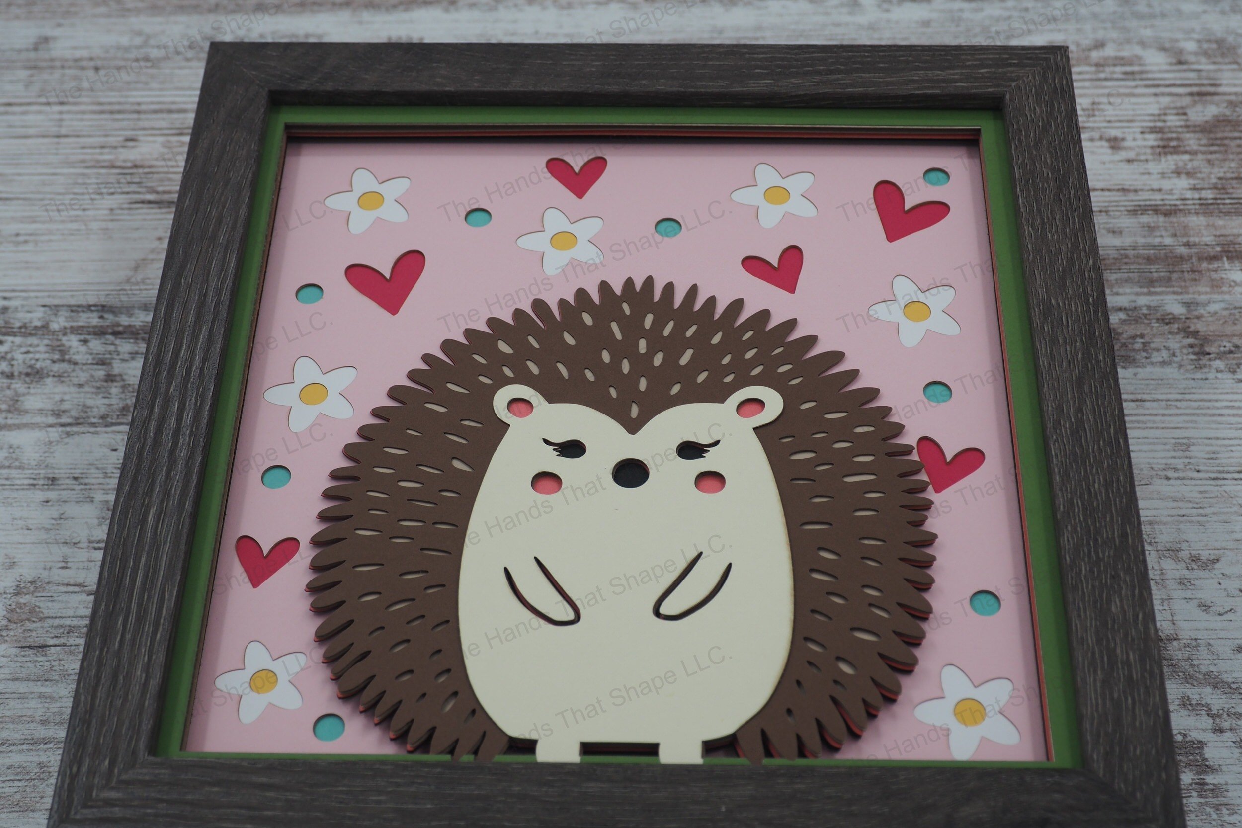 This layered paper shadowbox features a whimsical hedgehog, surrounded by flowers, dots, and hearts, perfect for adding a touch of folksy charm to your home.