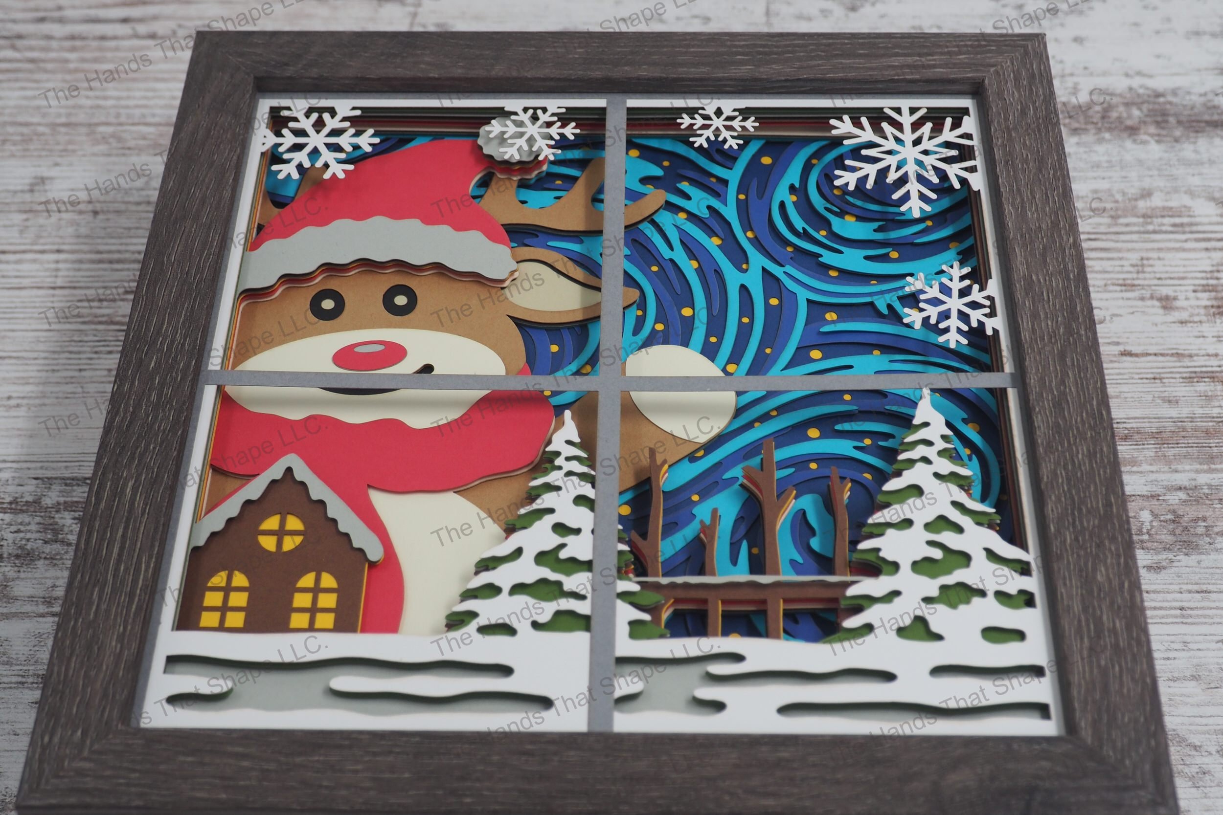 A layered paper shadowbox depicting an oversized festive reindeer through a window waving joyfully, with wintery swirls and stars as a backdrop.
