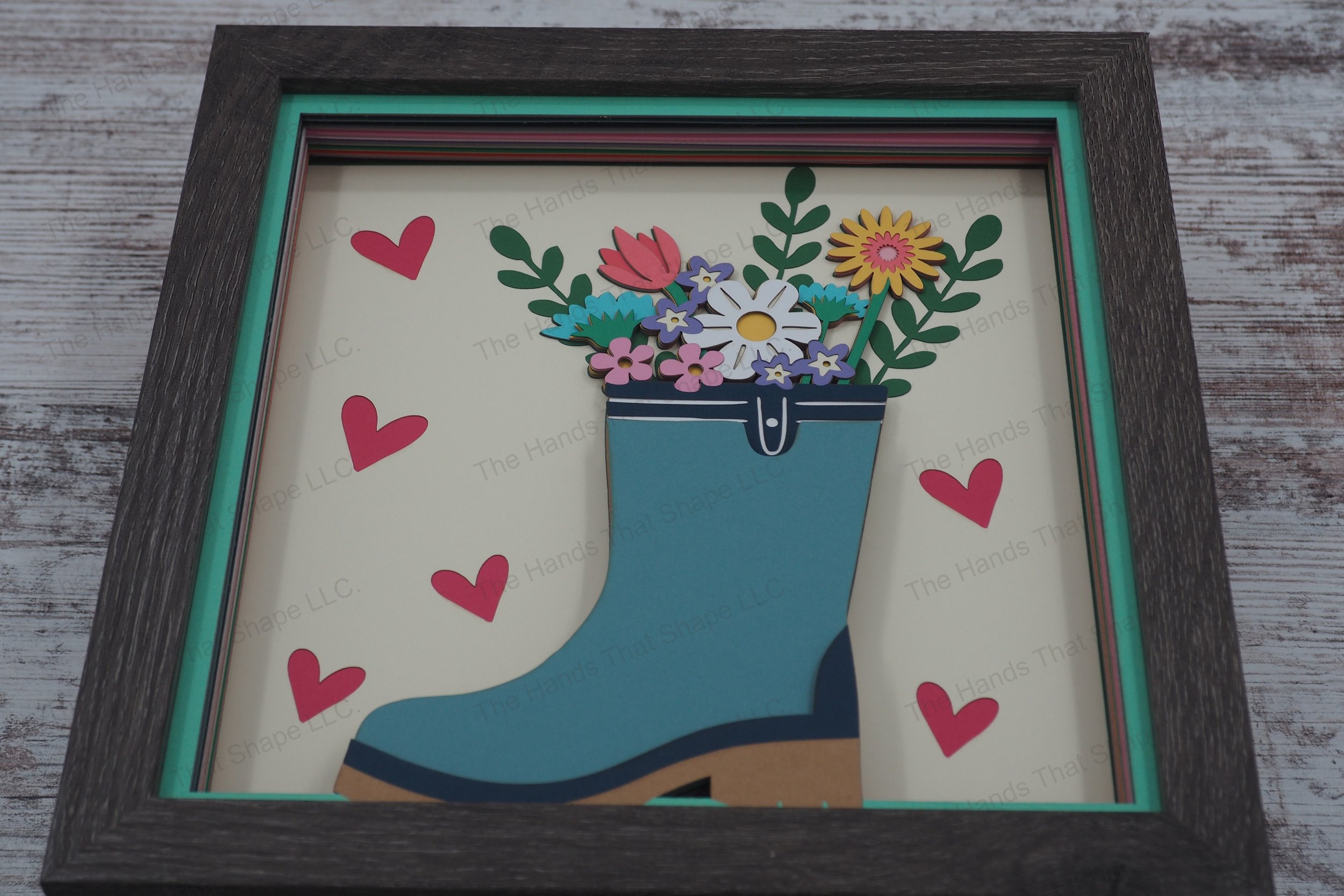 This springtime layered paper shadowbox features a bouquet of daisies, straw flowers, and lush greenery arranged playfully in a whimsical rain boot vase.