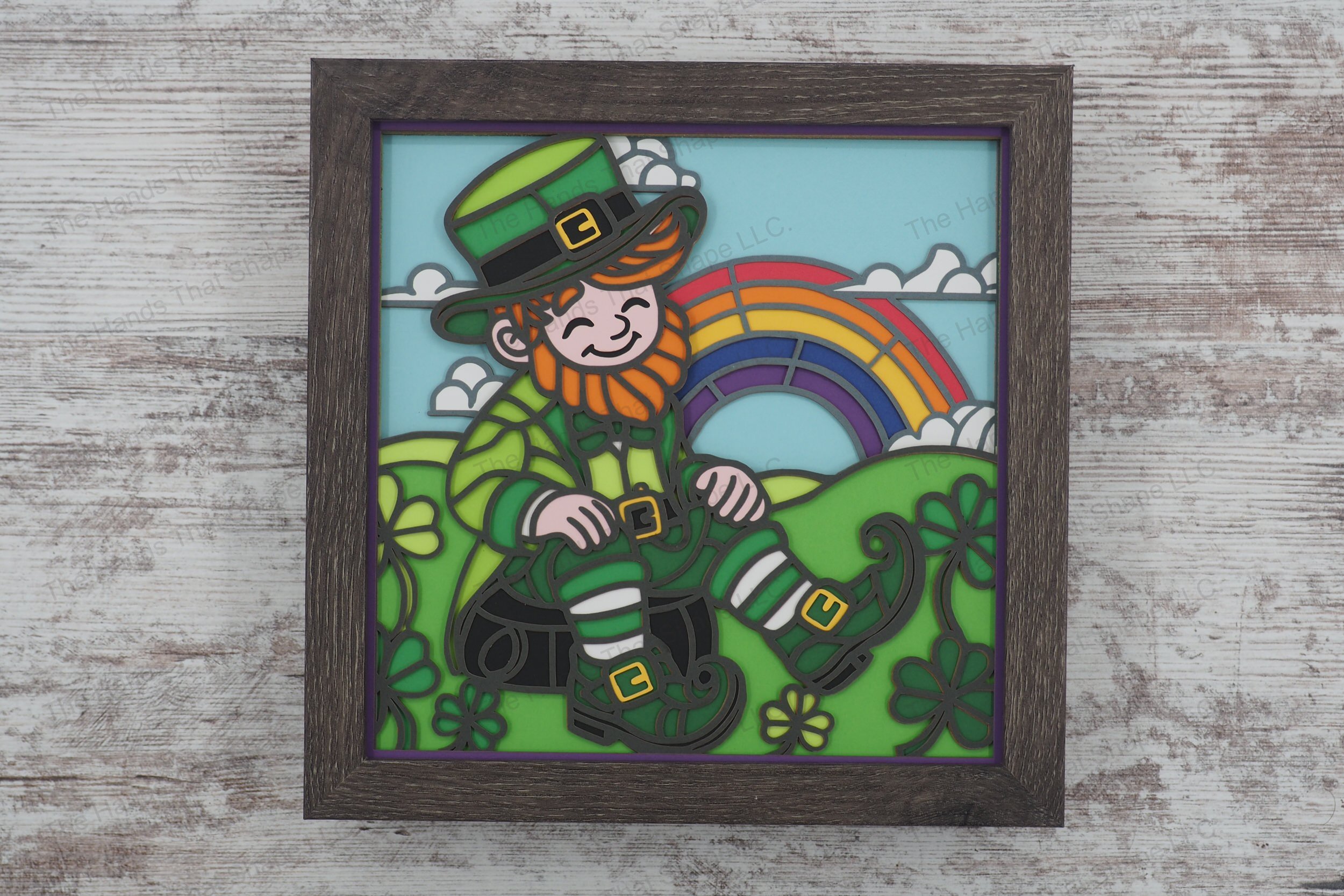 A joyful leprechaun sits atop a pot of gold, surrounded by shamrocks and rolling hills beneath a vibrant rainbow in this intricately layered paper shadowbox.