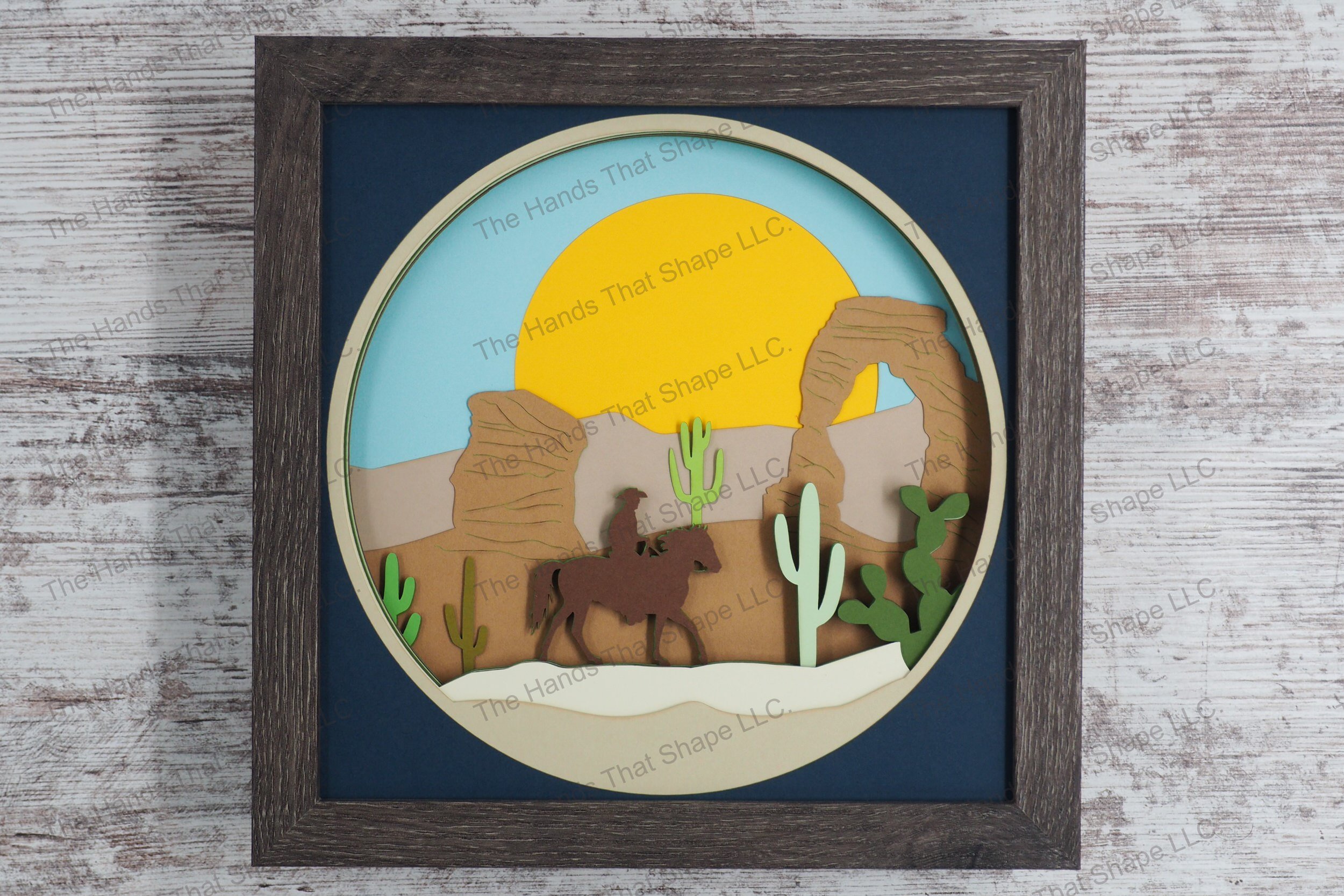 A striking depiction of a cowboy on horseback navigating through a desert landscape, this layered paper shadowbox features cacti, a stone arch, and sun.