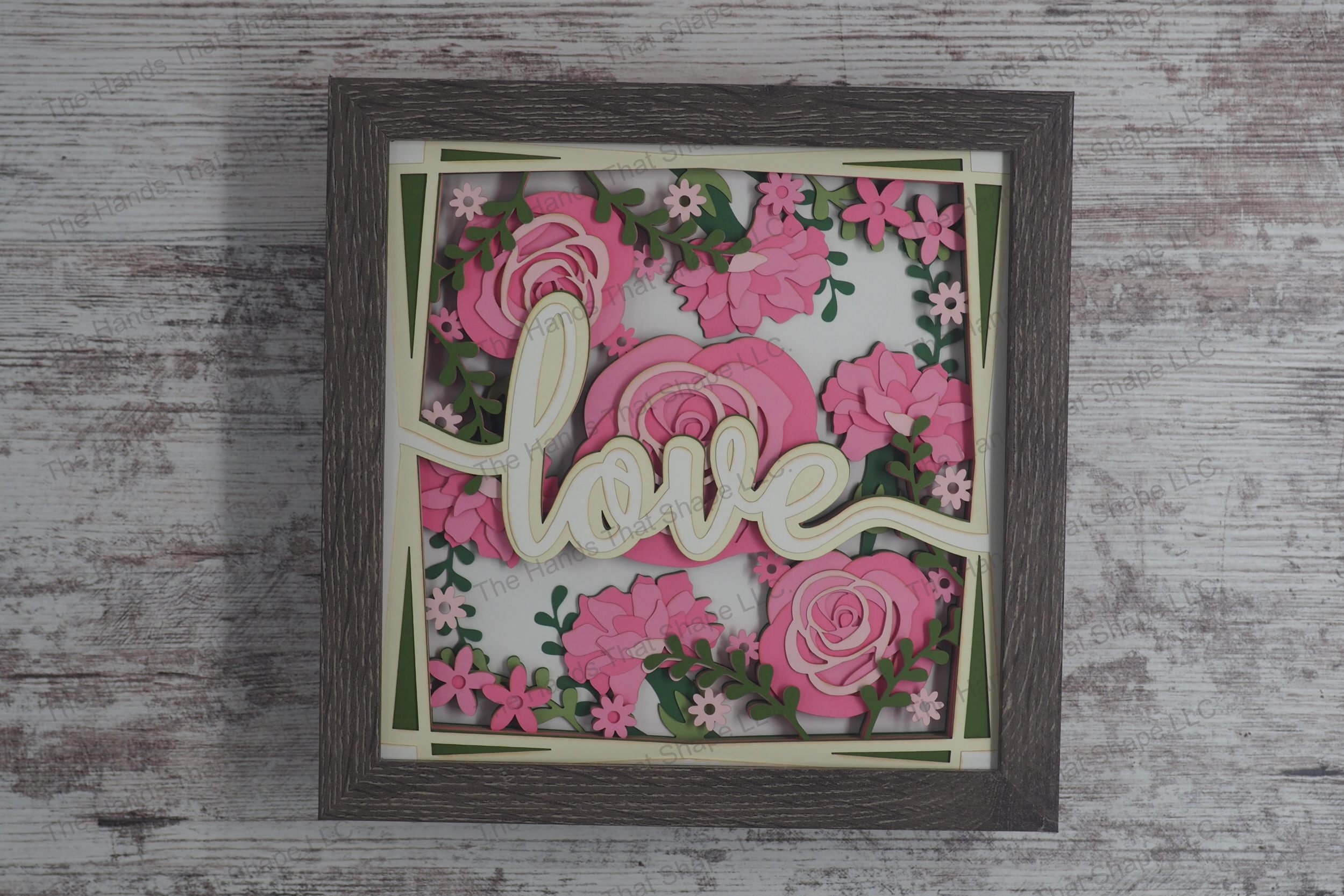A romantic layered paper shadowbox featuring the word 'Love' surrounded by vibrant, multi-layered roses and greenery, crafted from cardstock in a gray frame.