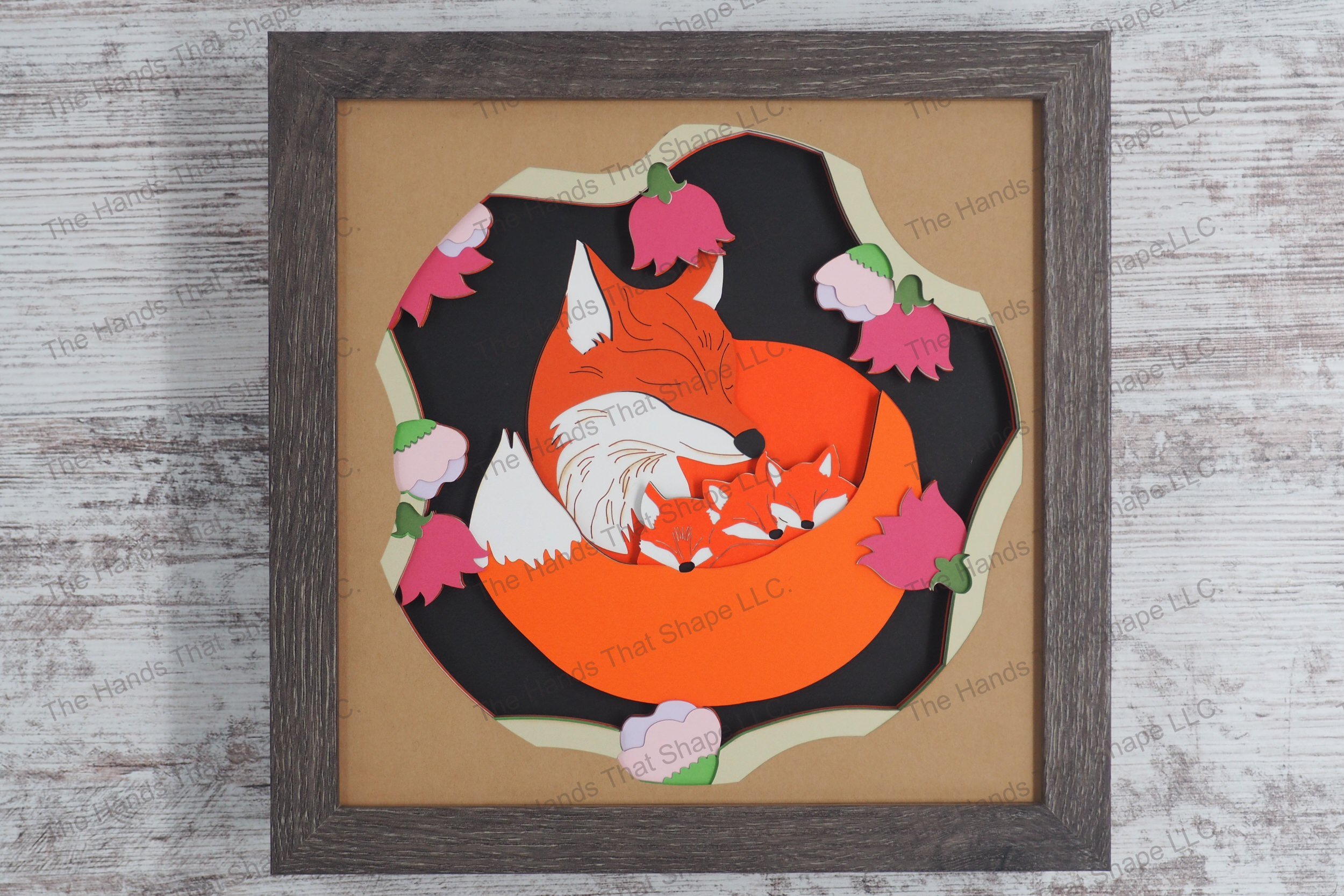 A captivating layered paper shadowbox depicting a mama fox tenderly curled around her three kits among vibrant flower blossoms in a gray MDF frame.