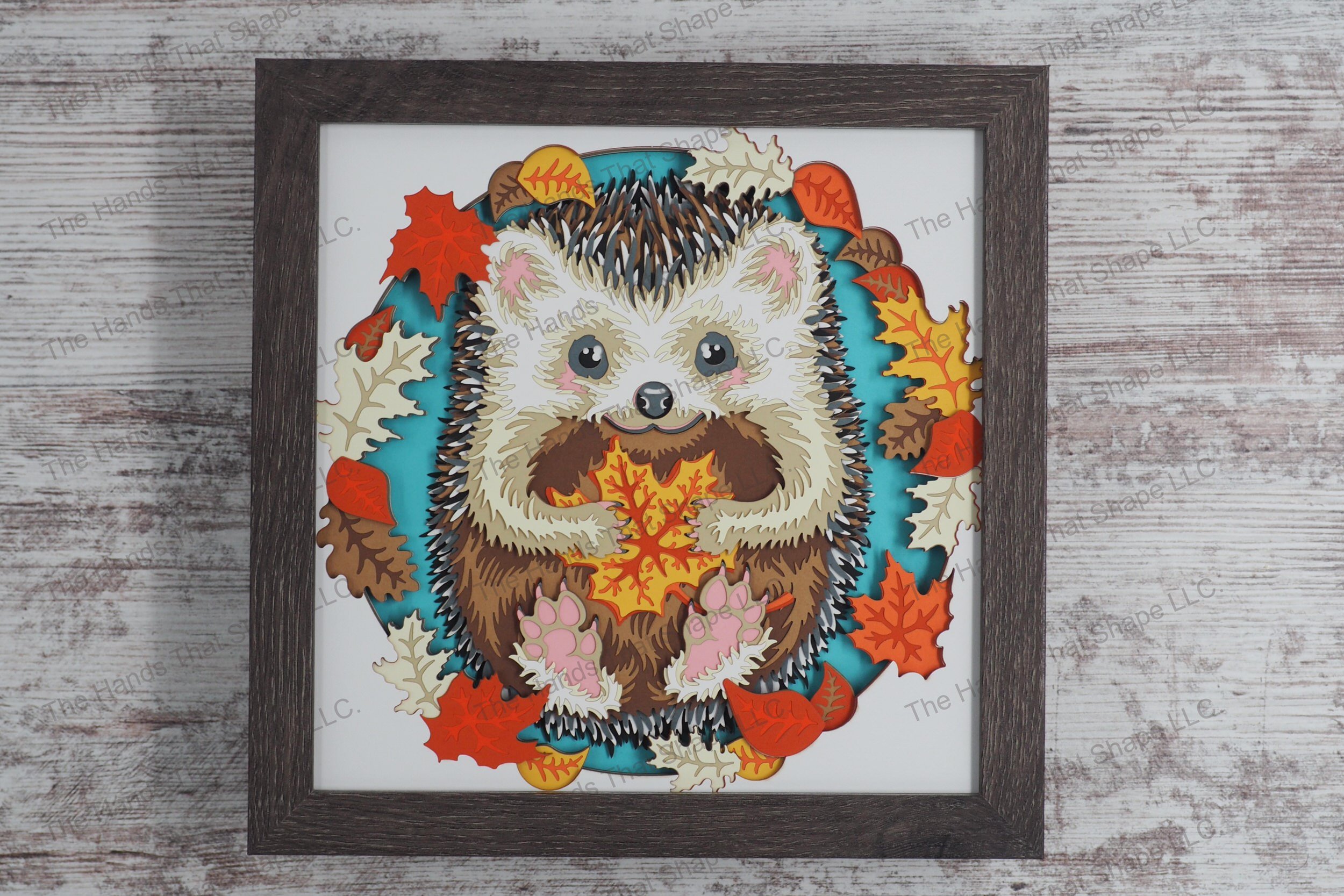A captivating layered paper framed shadowbox depicting a hedgehog clutching a colorful autumn leaf, surrounded by a frame of fall foliage.