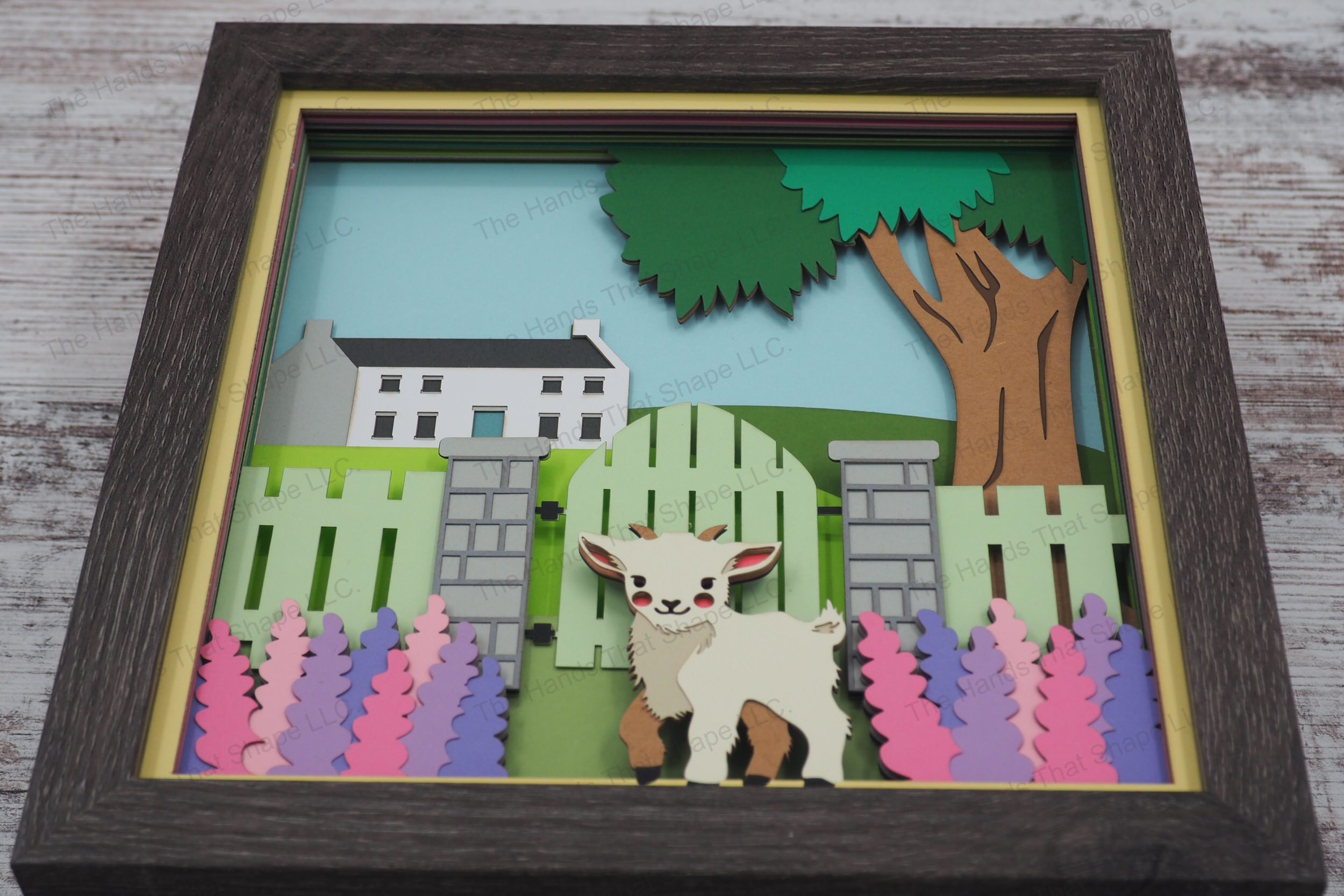 This layered paper art shadowbox features a goat standing in a flower garden, beside a rustic fence framed by blooming hyacinths and a countryside house.