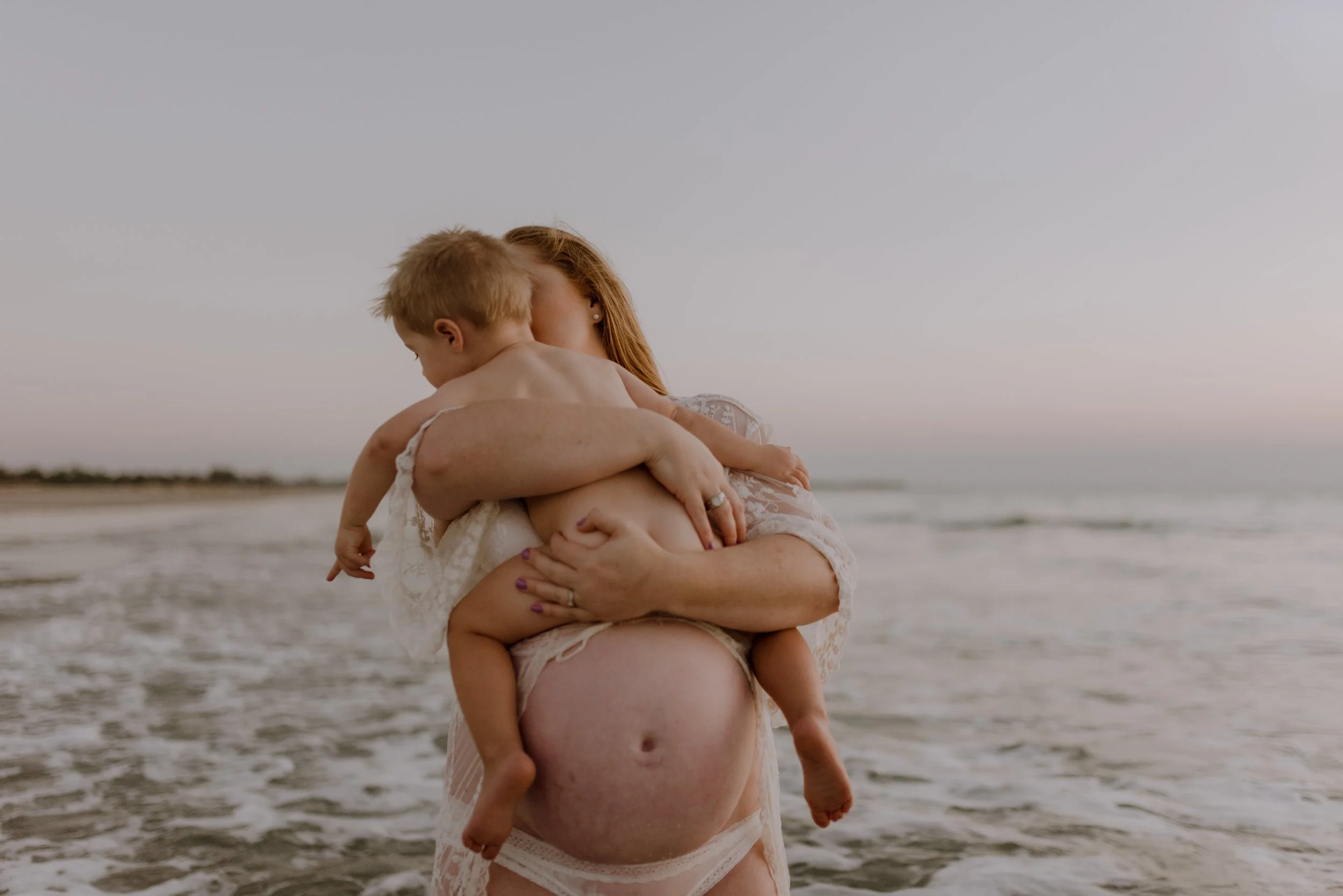 A woman holding a child on a beach at sunset, with the ocean and sky in the background.