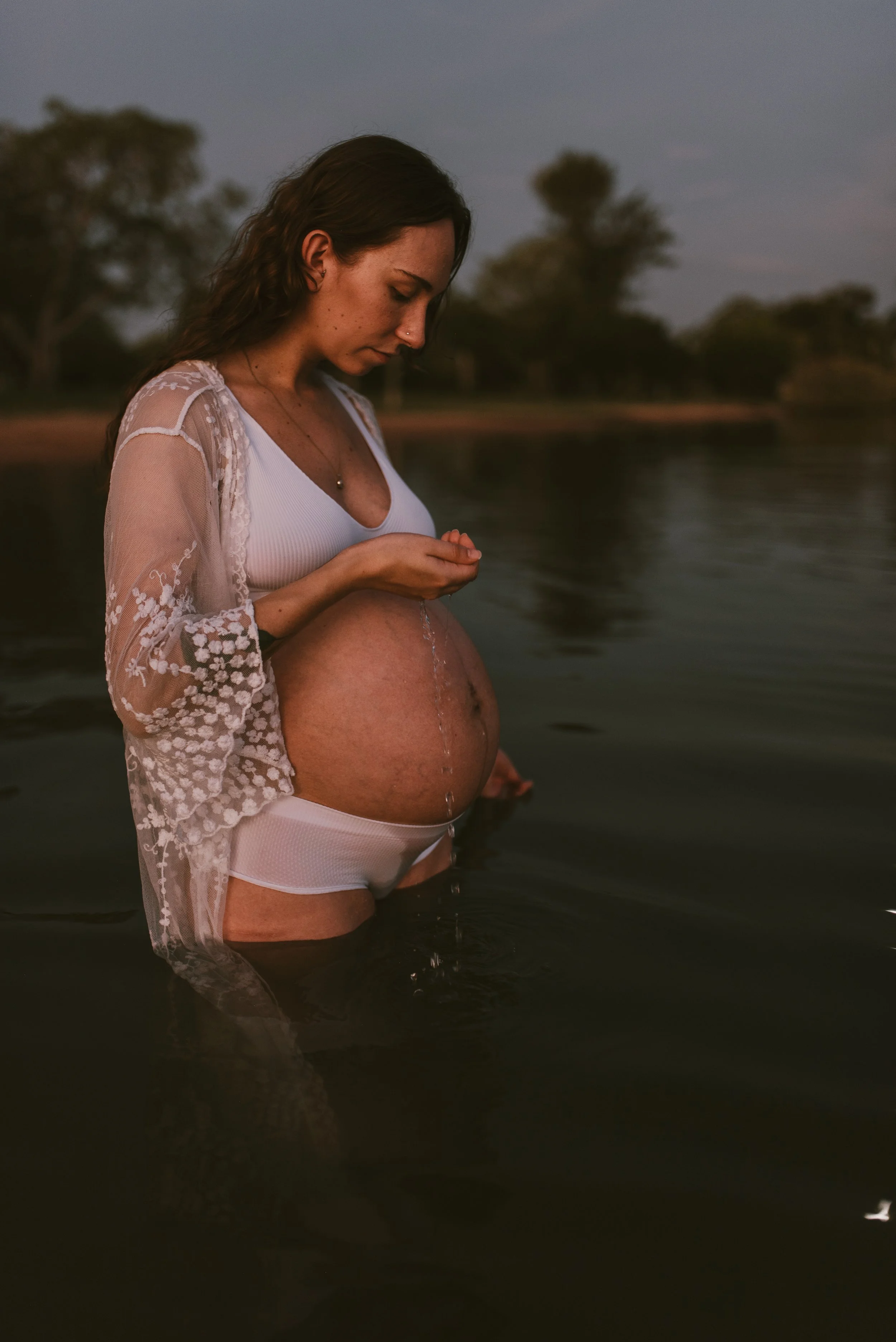 A pregnant woman standing in water at dusk, wearing white lingerie and a sheer embroidered cover-up, with trees in the background.
