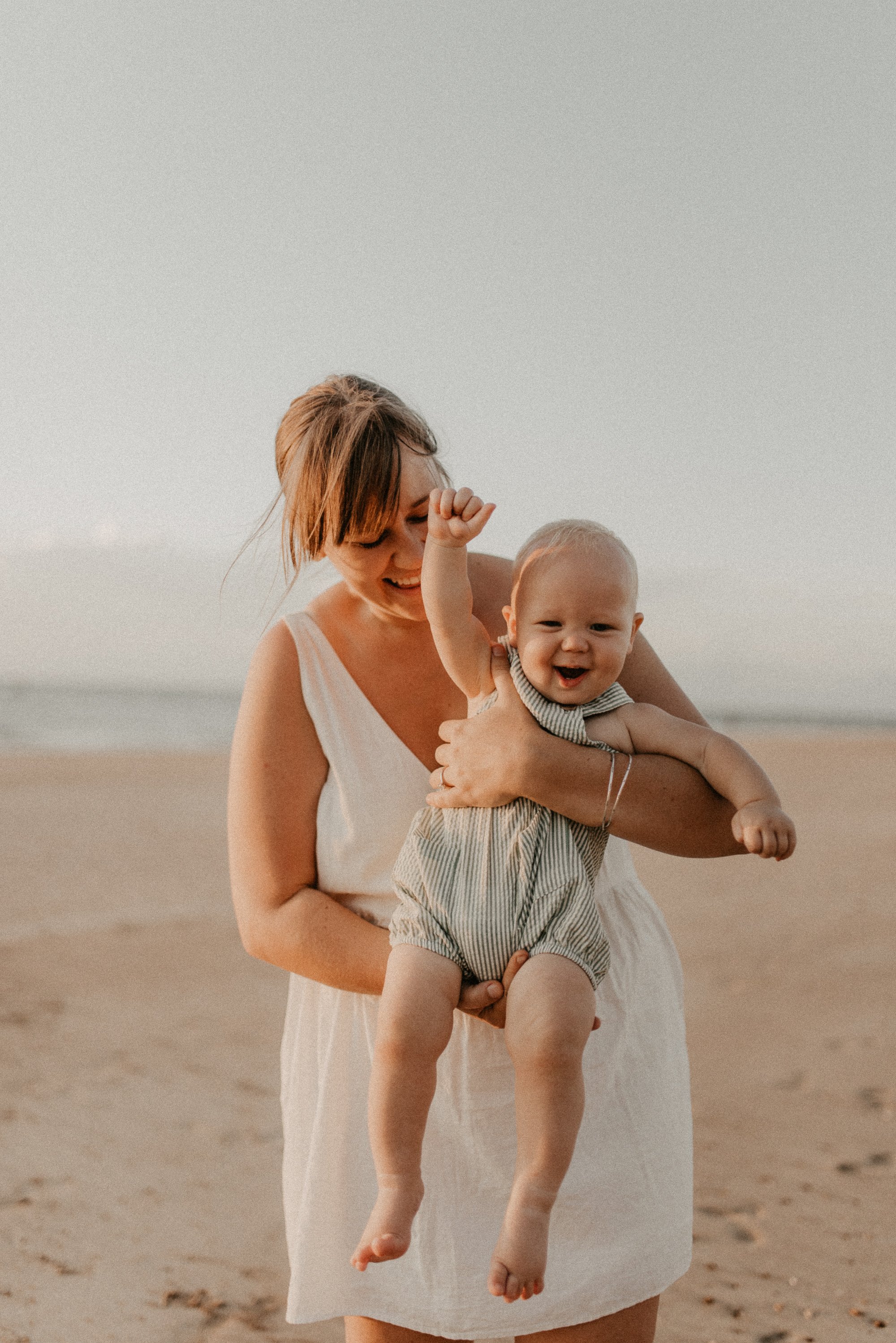 A woman holding a smiling baby at the beach during sunset, with the ocean in the background.