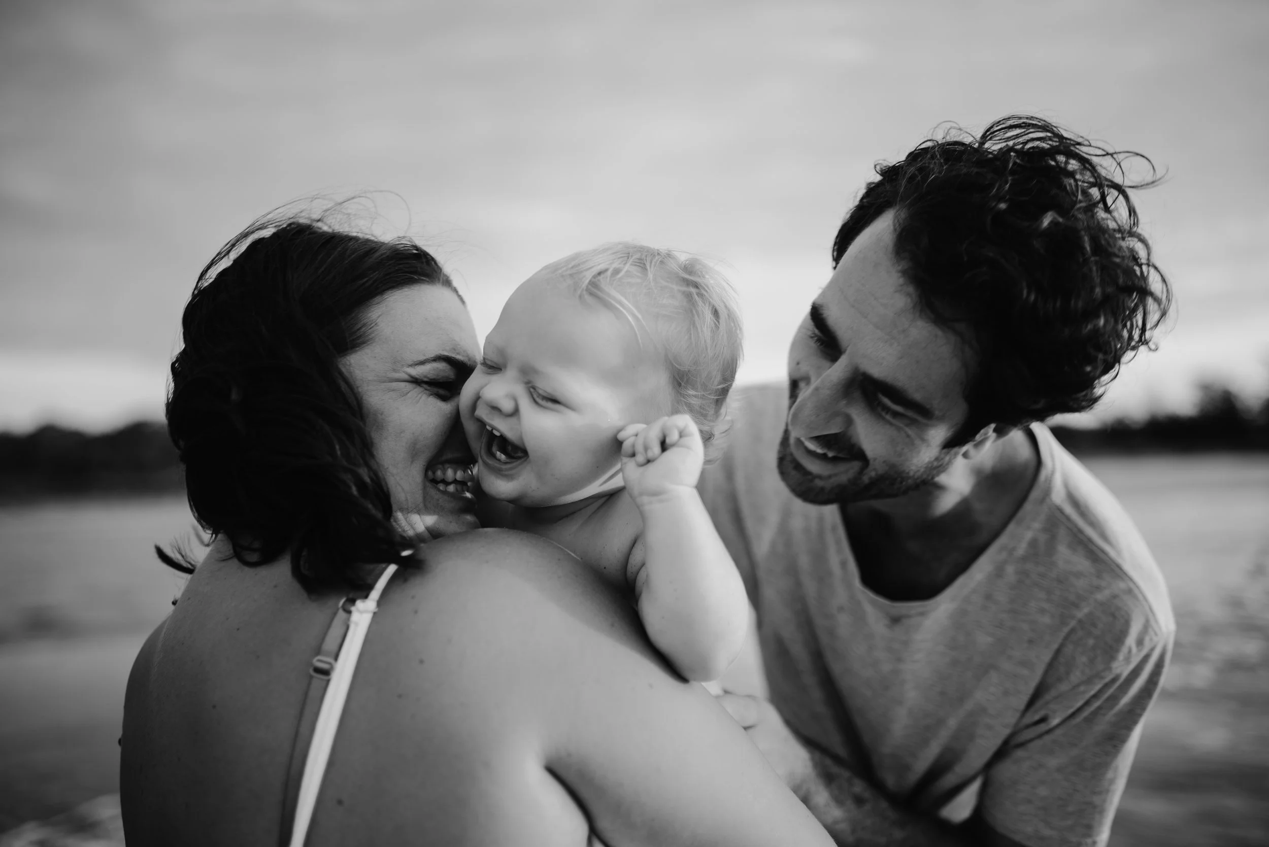 A black and white photo of a happy family of three enjoying time together at the beach, with the mother holding a smiling toddler and the father leaning in close.