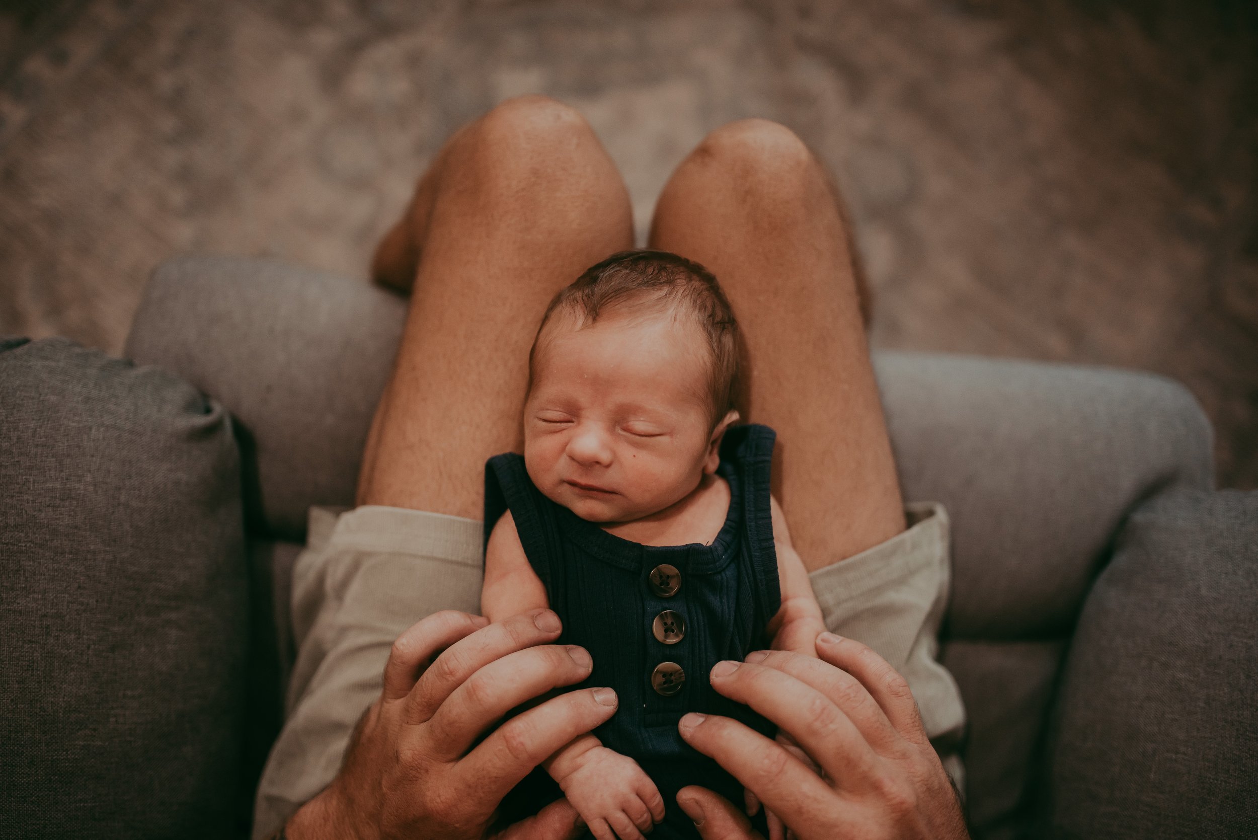 A newborn baby resting on a person's lap, with eyes closed and a slight smile, wearing a dark sleeveless shirt with buttons. The person's legs are visible from above, and the background shows a wooden floor.