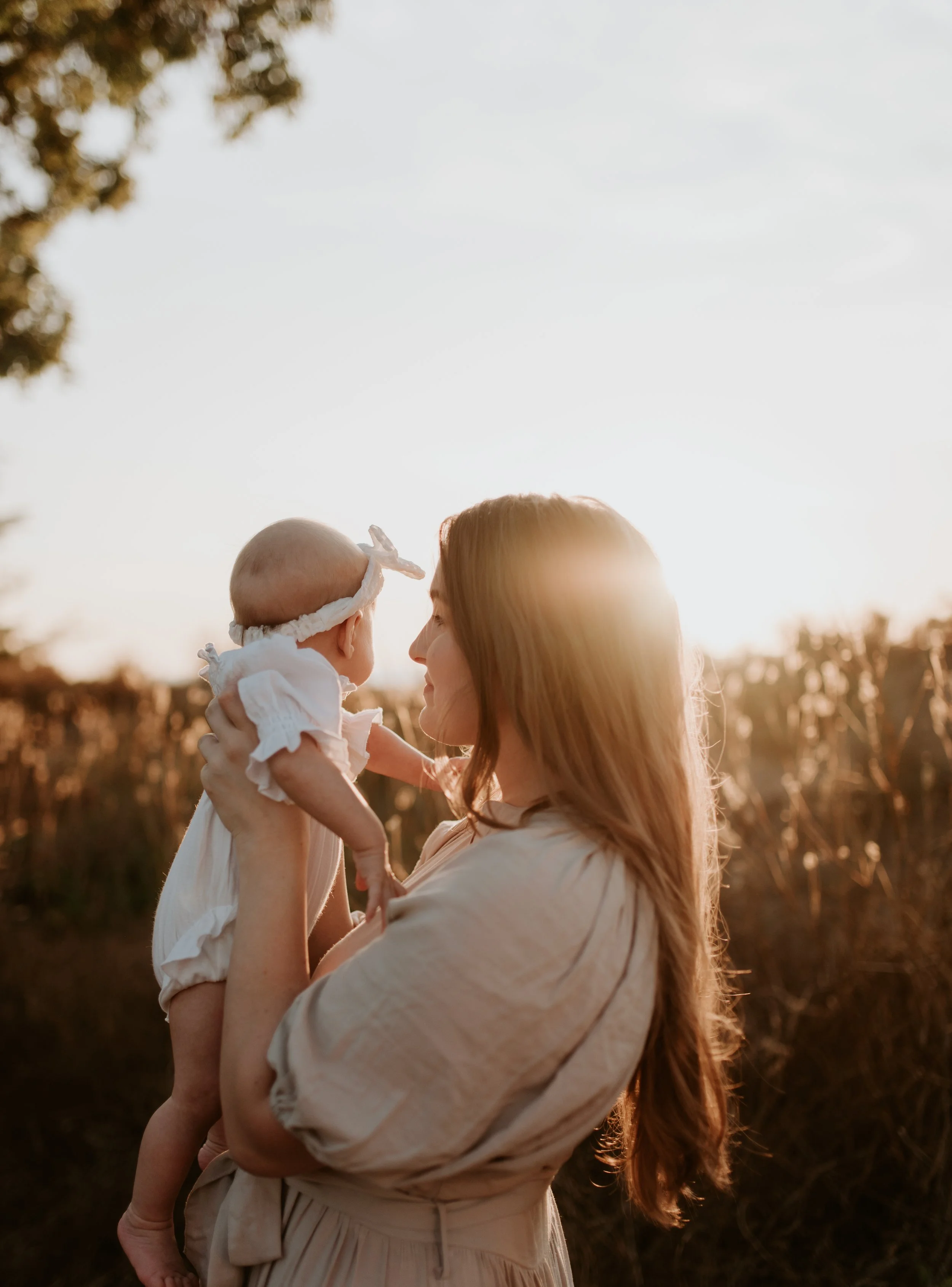 A woman holding a baby girl outdoors during sunset, with trees and a clear sky in the background.