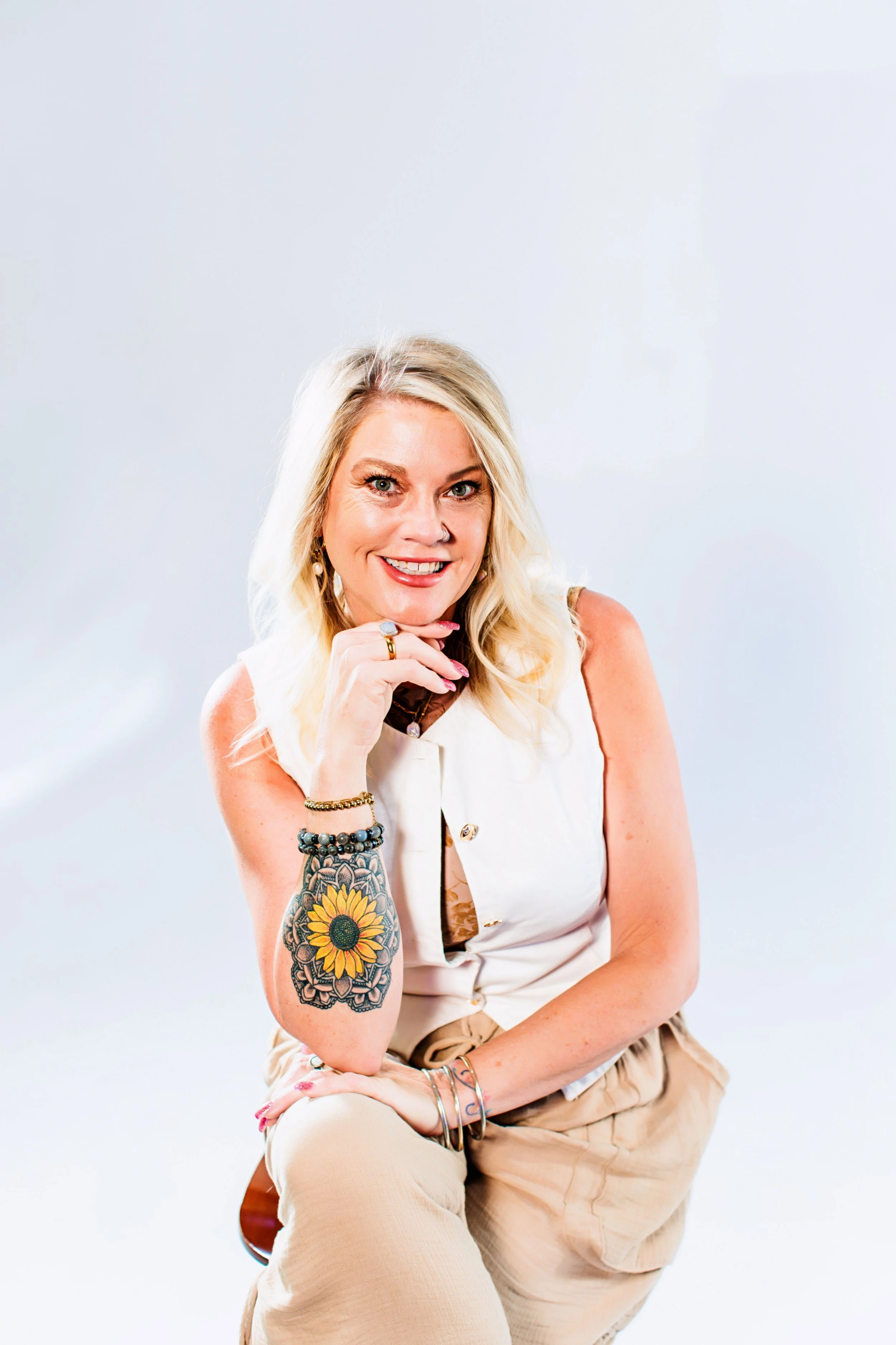 A woman with blonde hair and tattoos, sitting on a chair, smiling with her hand near her chin, against a plain white background.
