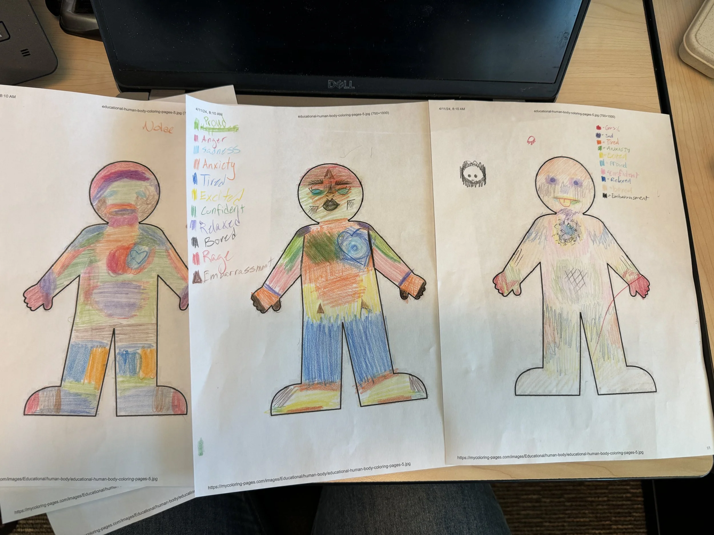 Three sheets of paper showing children's colorful crayon drawings of human figures, each with different emotions and features, accompanied by a color-coded list of emotions on the left side.