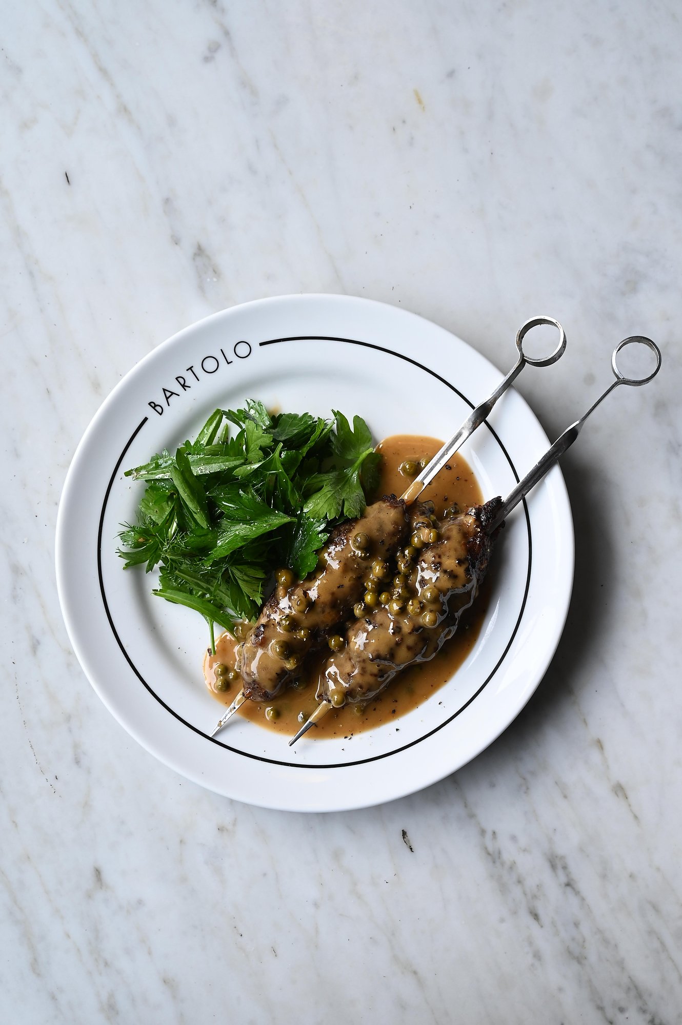 A white plate with the words 'BARTOLO' on the rim, holding a piece of meat on a skewer topped with peppercorn sauce, served with fresh green herbs and watercress, on a marble surface.