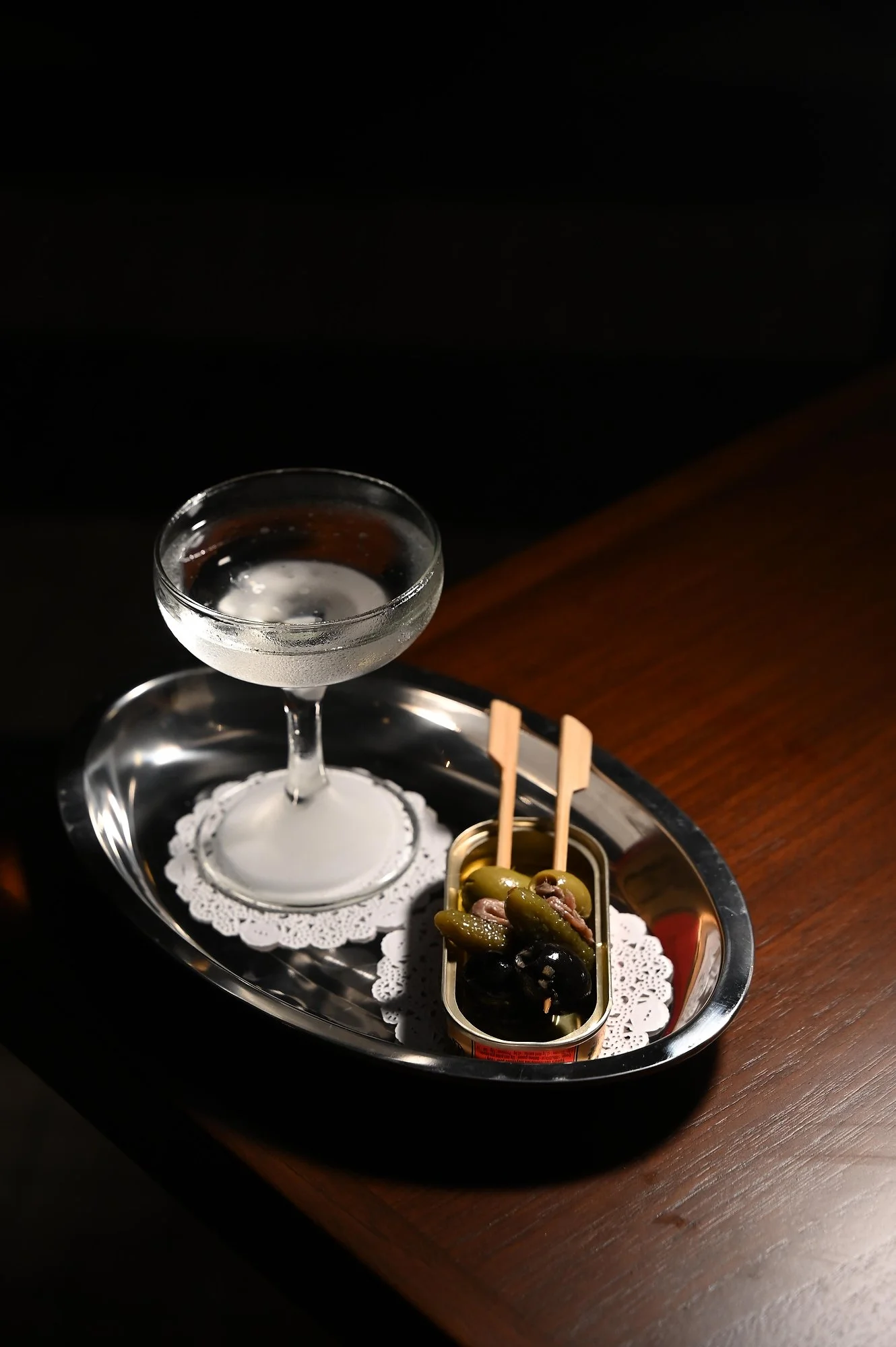 A glass of clear beverage on a tray with olives and pickles, placed on a dark wooden table.