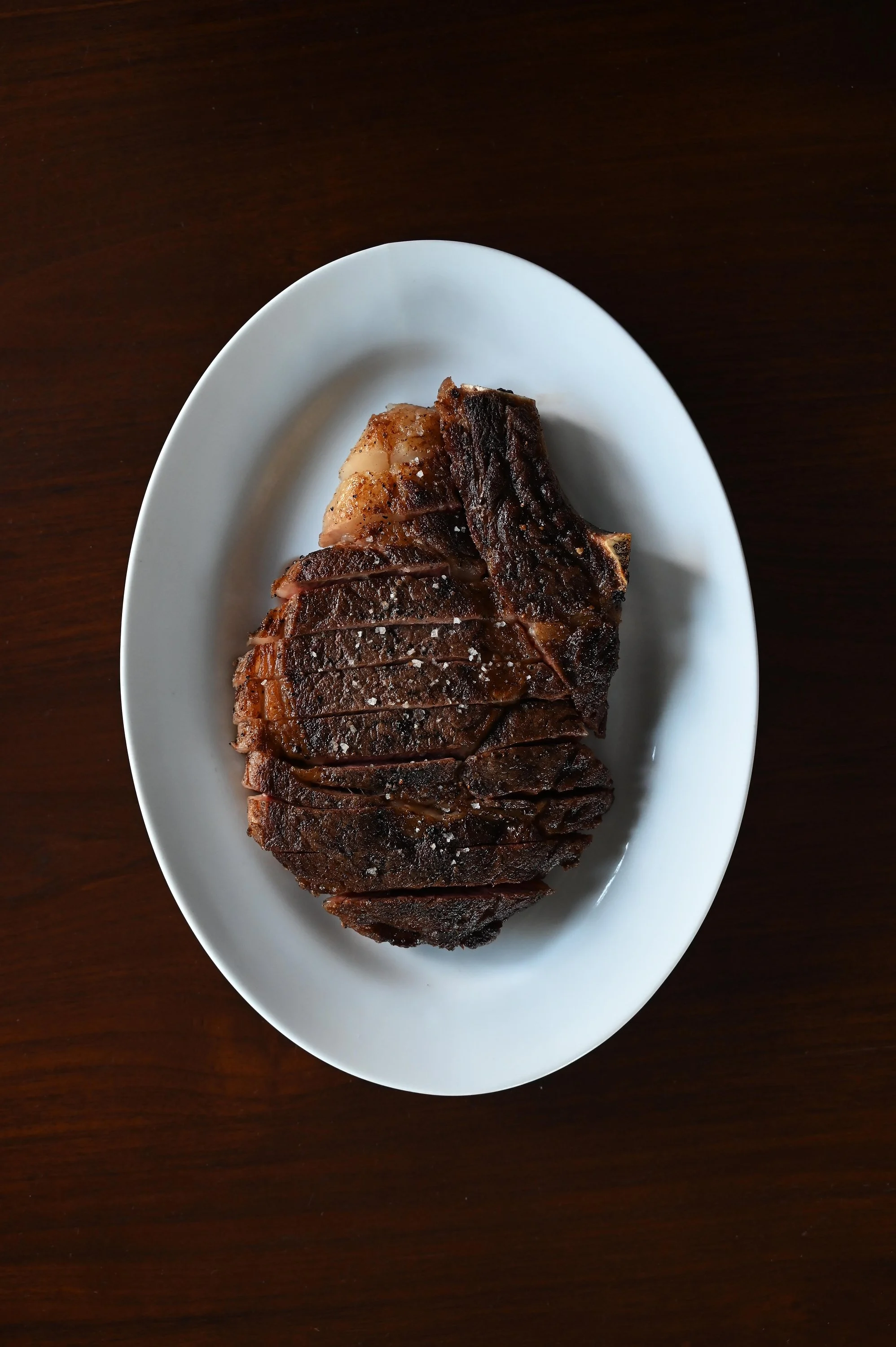A white oval plate with a cooked ribeye steak, seasoned with salt, on a dark wooden table.