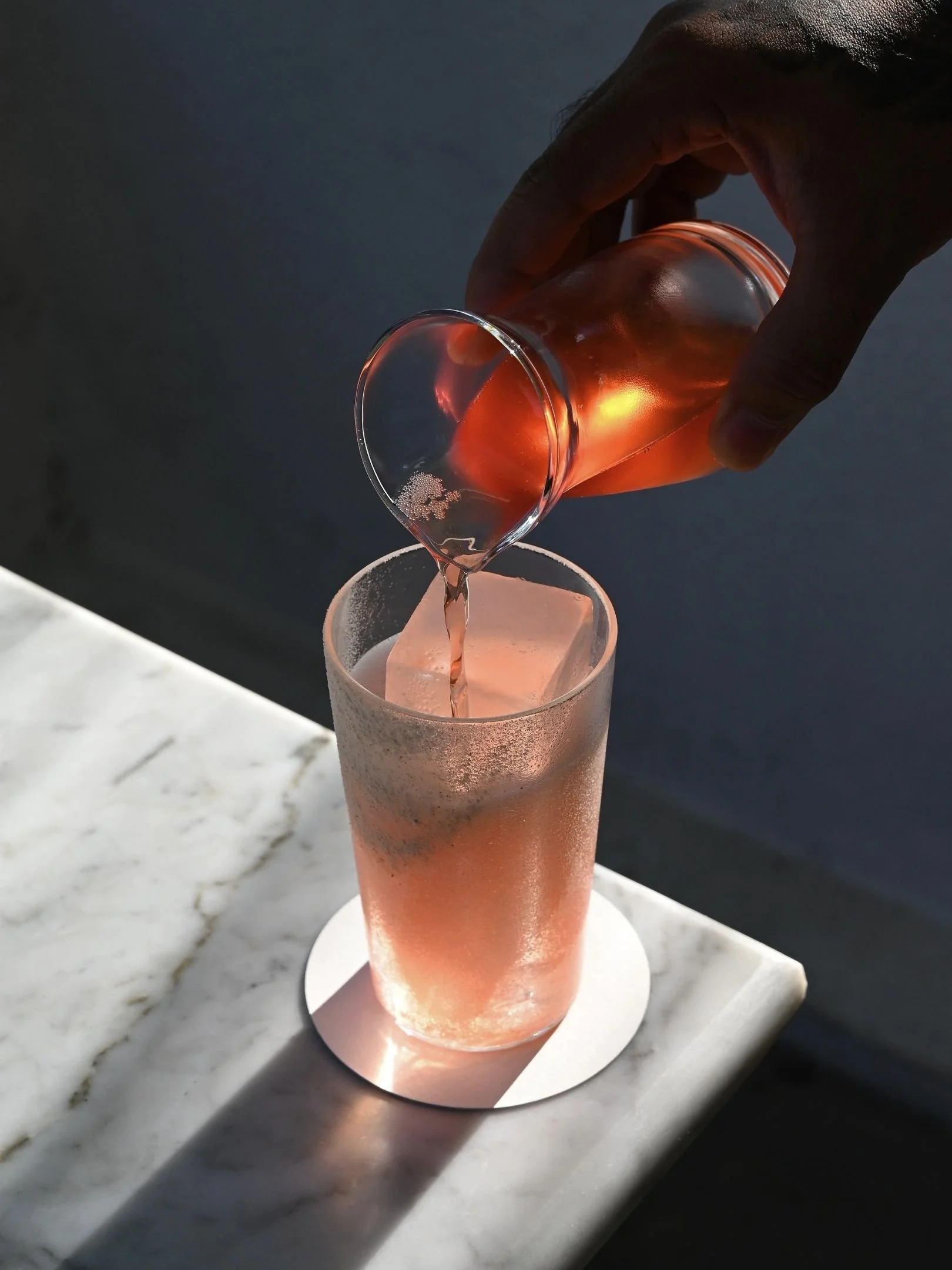 A hand pouring a pinkish drink into a tall glass with ice on a marble surface, with a dark background.